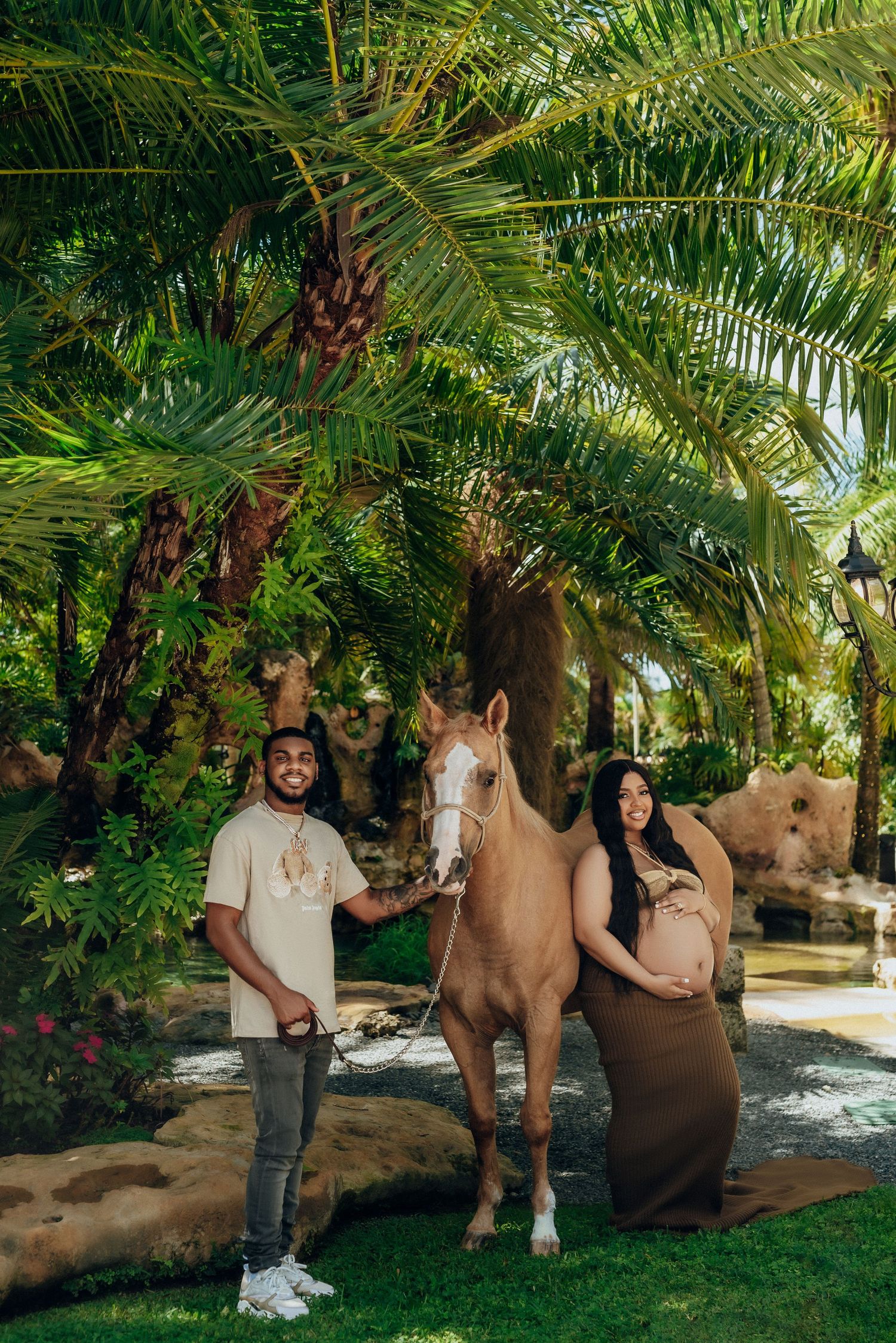 A brown horse stands between two people under lush palm trees in a tropical garden setting.