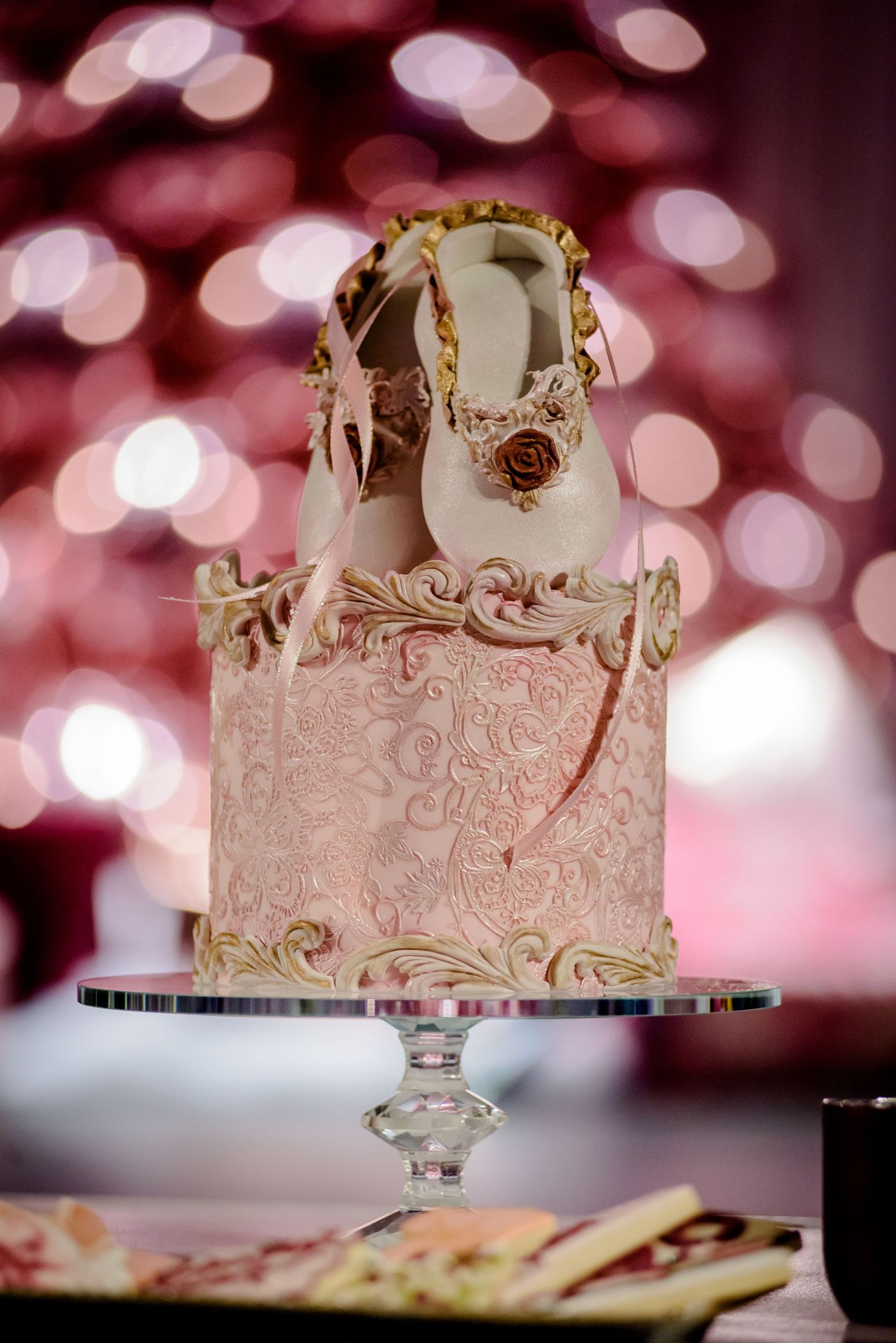 Elegant pink and gold wedding cake with ornate details and bokeh lighting in the background.