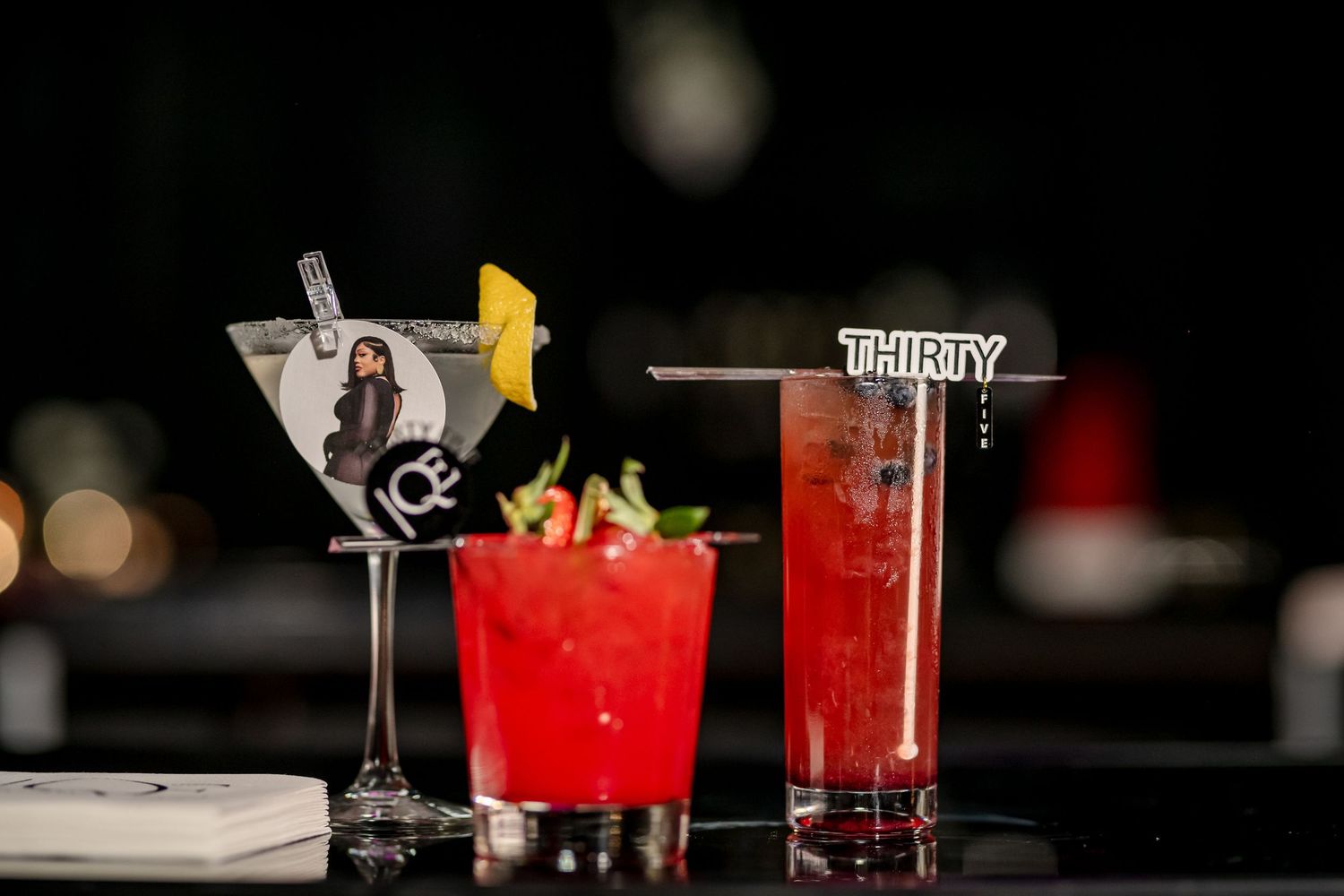 Red cocktails with garnishes are displayed in elegant glassware on a dark bar counter with ambient lighting.