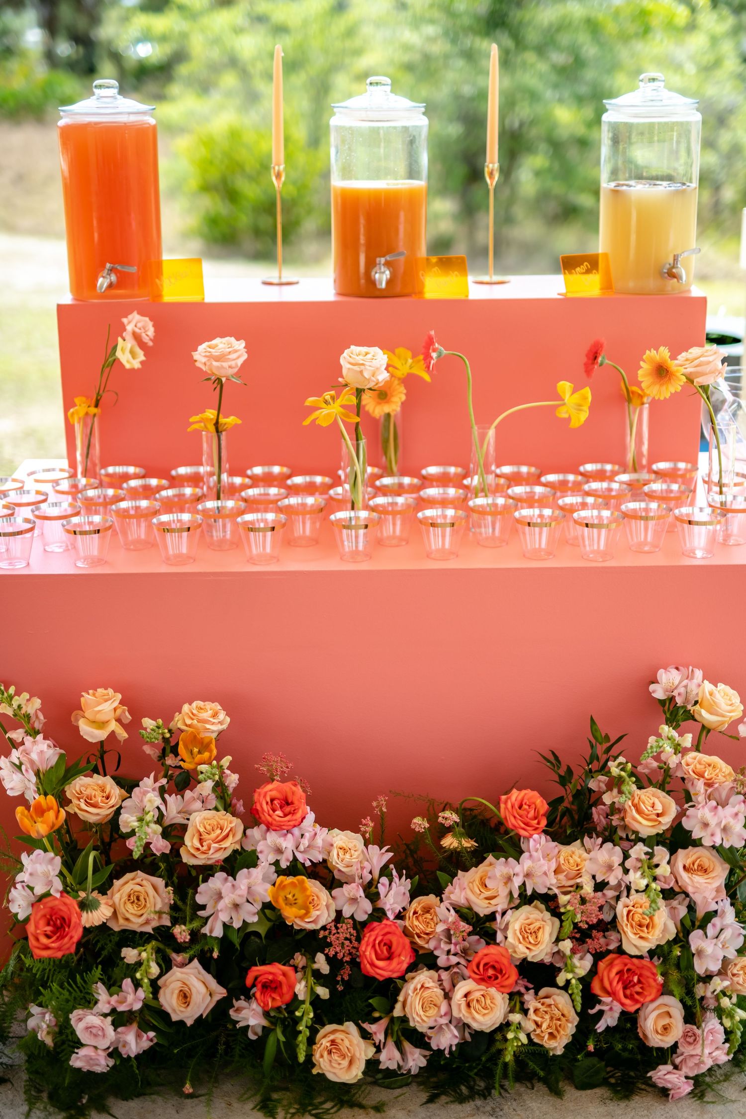 A pink drink station decorated with coral and peach roses features three beverage dispensers filled with colorful juices.