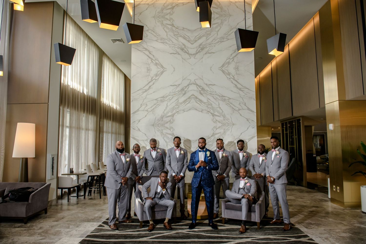Groomsmen in light gray suits pose confidently against modern marble wall backdrop.