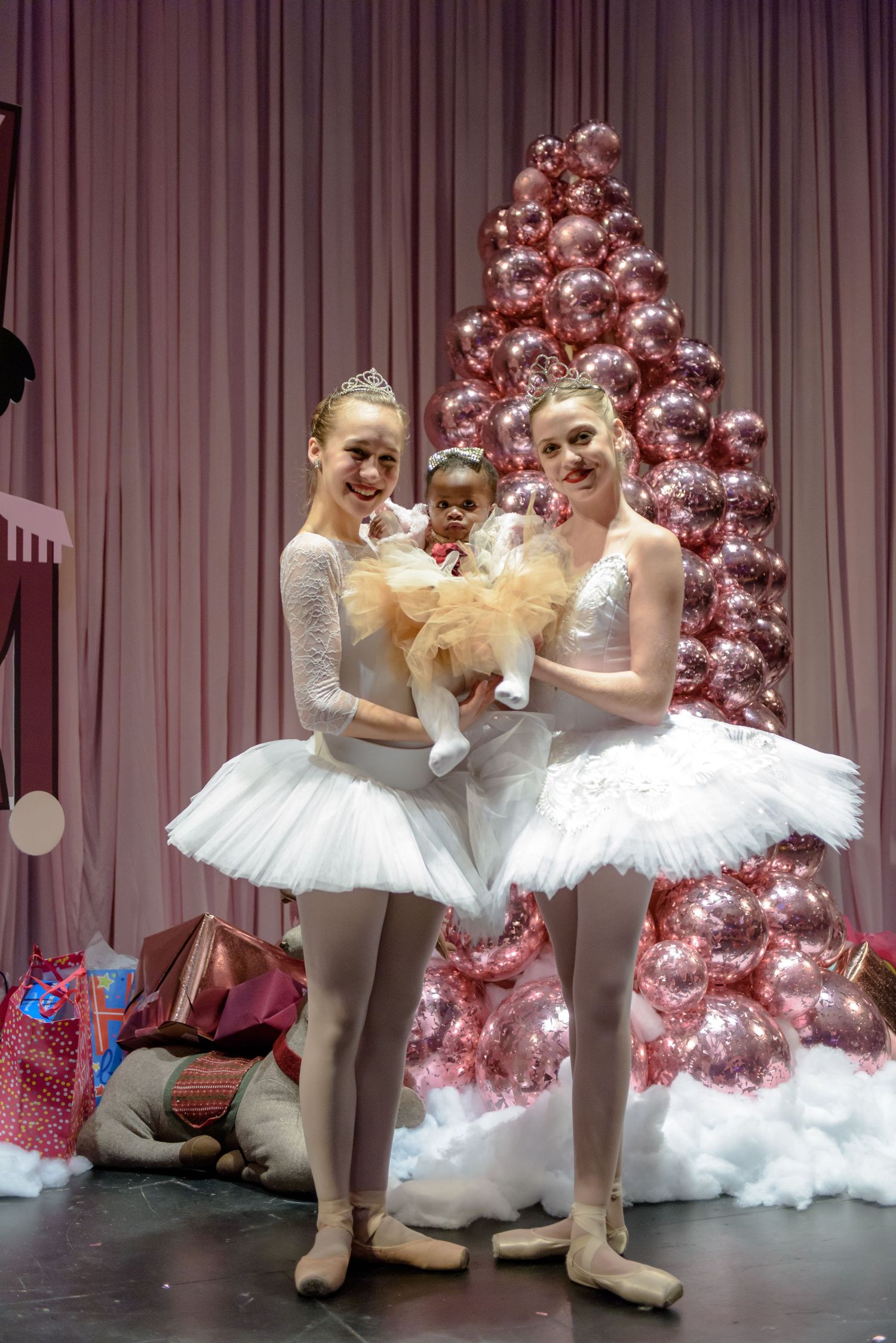 Two ballet dancers in white tutus pose in front of a pink Christmas tree on stage during a holiday performance.