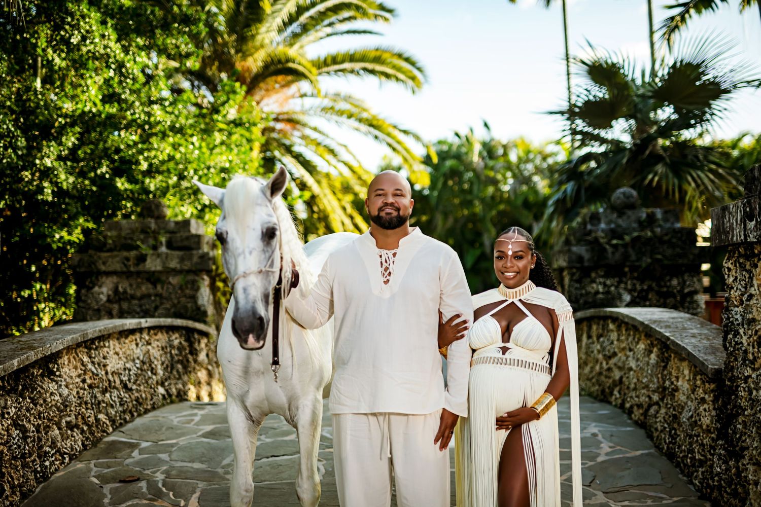 Wedding party in white attire walking along a palm-lined stone path outdoors.