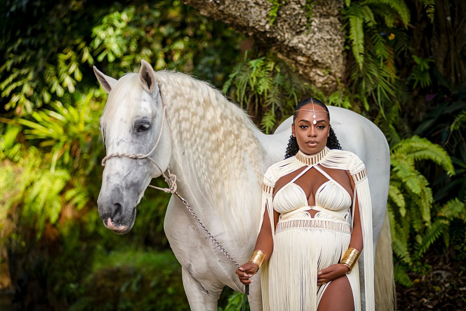 A white horse stands next to someone in a white dress against a lush green garden backdrop.