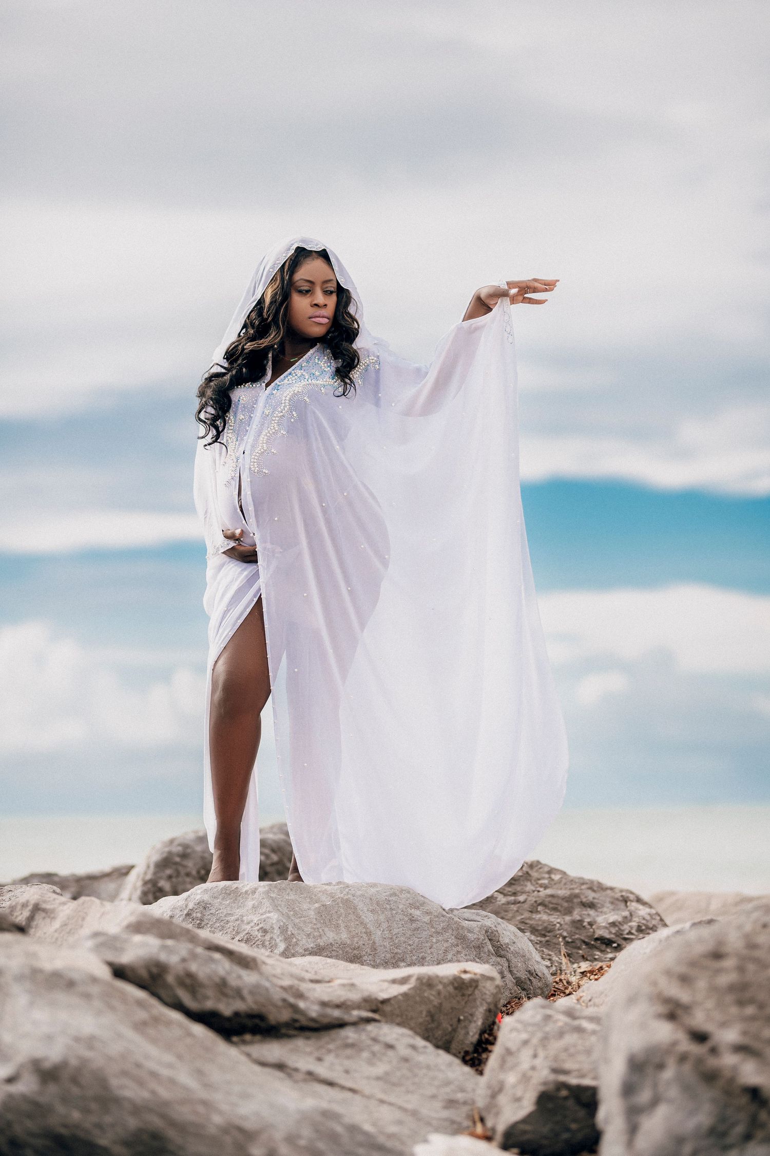 Person in flowing white garments poses dramatically on rocky terrain against a blue sky with clouds.