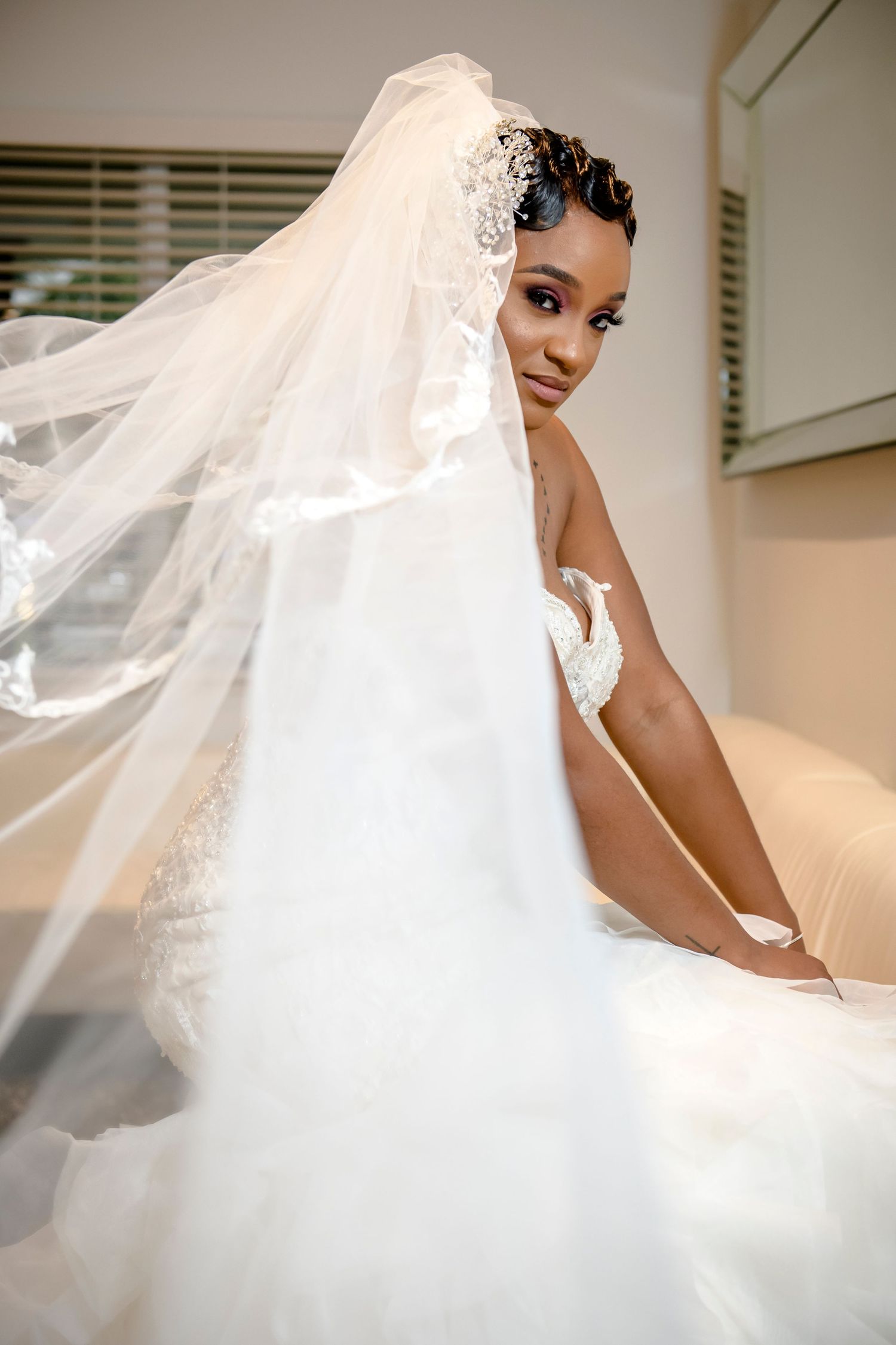 Elegant bride in white wedding dress and flowing veil poses gracefully in indoor setting.