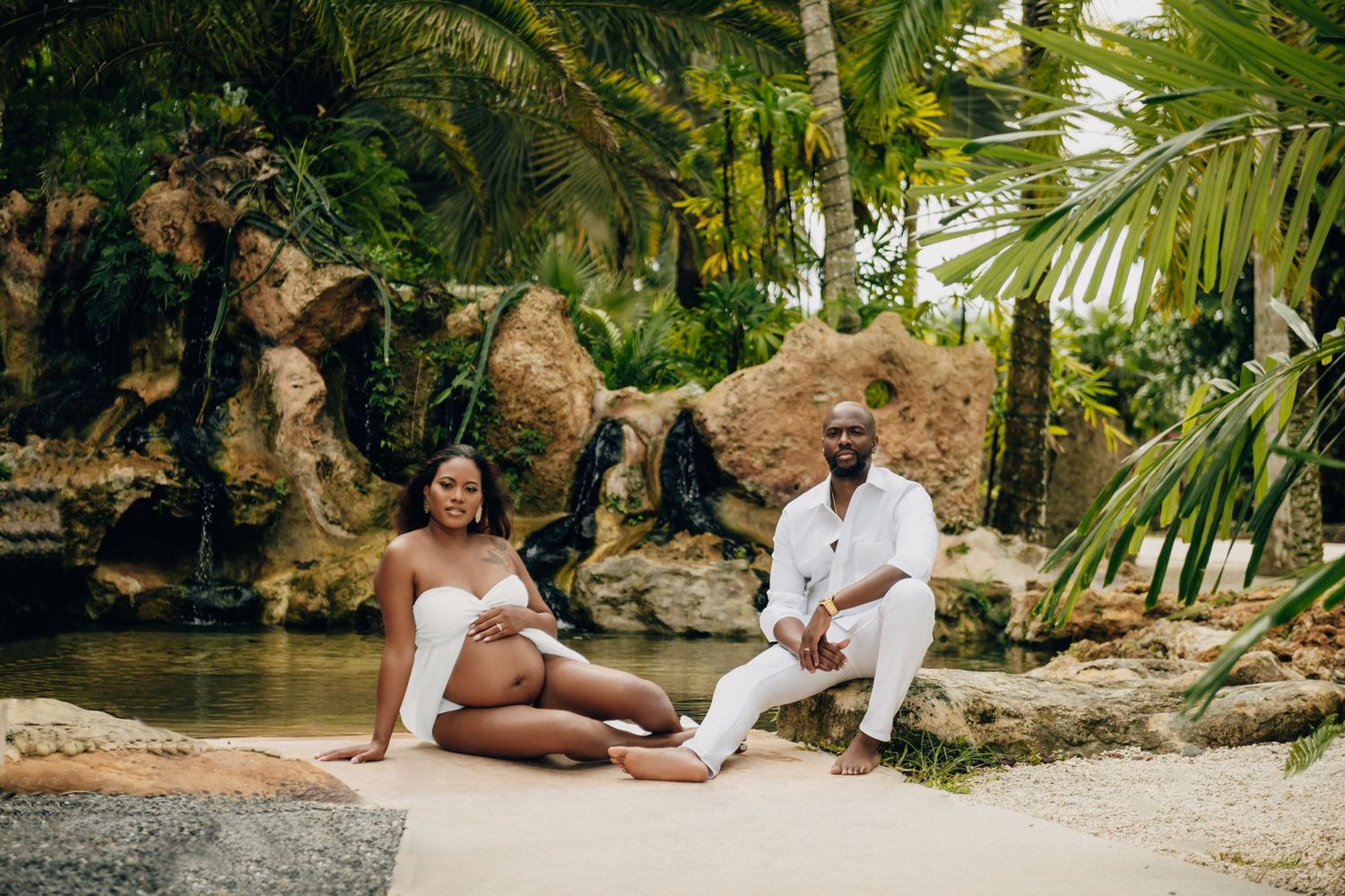 A couple in white outfits poses together in a lush tropical garden setting for romantic photos.