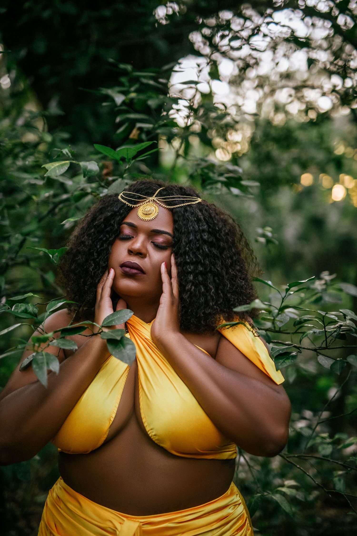 A person in a yellow dress poses gracefully among green foliage at sunset with a gold headpiece.