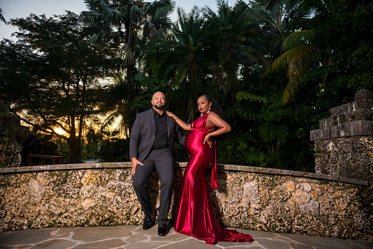 Couple in formal wear posing along a stone wall path at sunset.