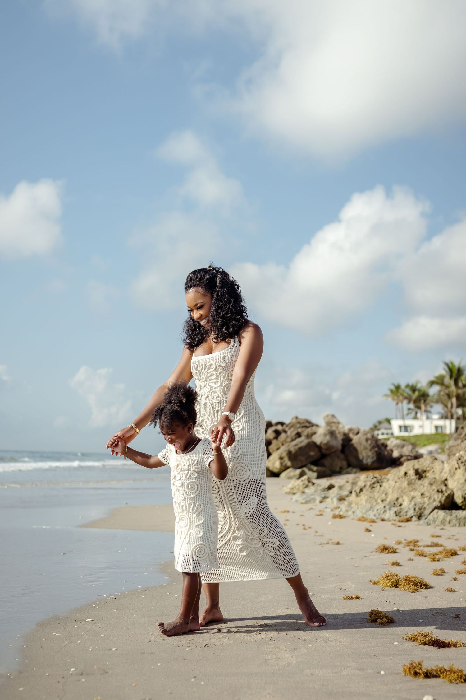 Two people in matching white lace dresses play together on a beach near rocky cliffs.