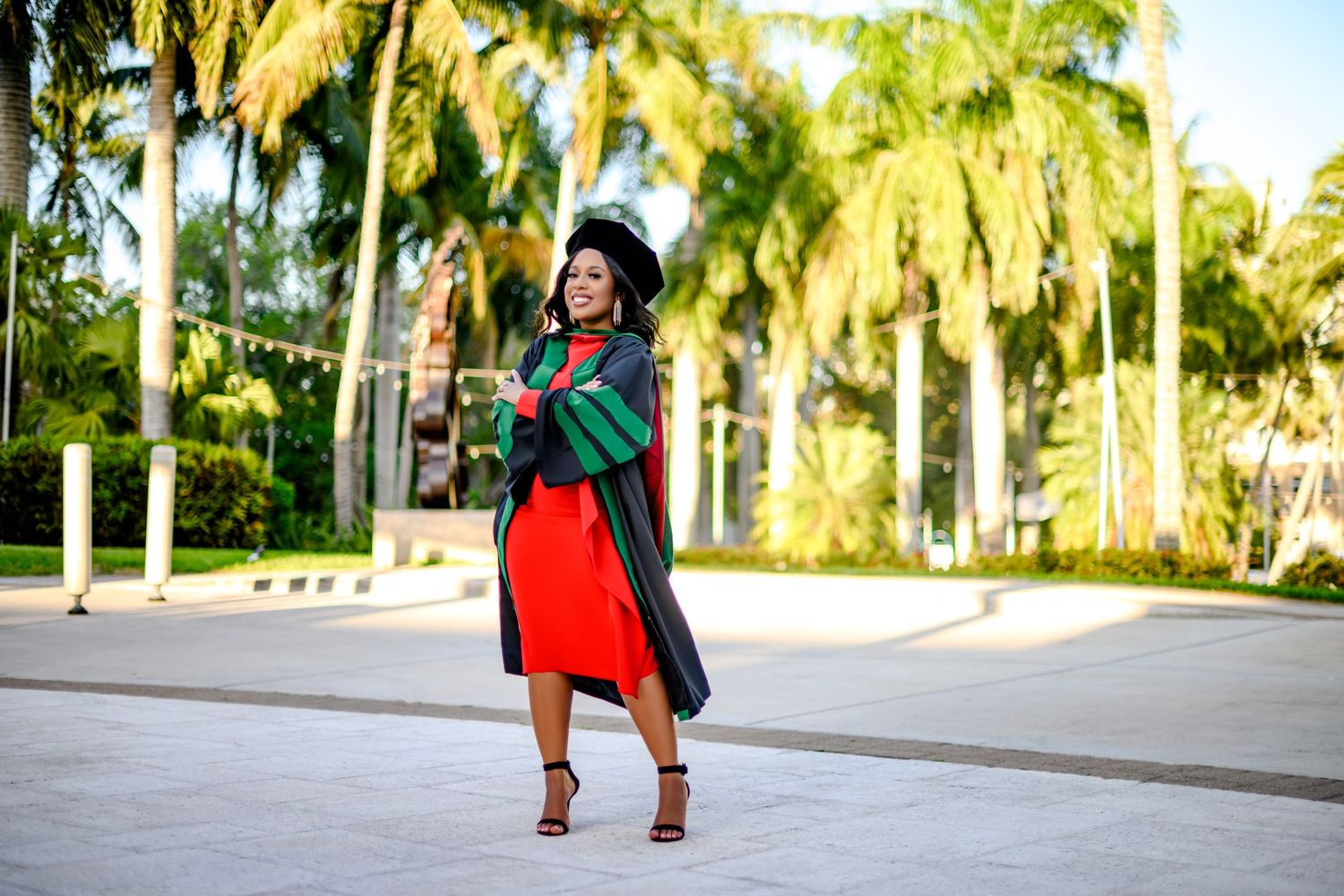 Fashionable graduate in red dress and green graduation gown poses outdoors with palm trees in background.