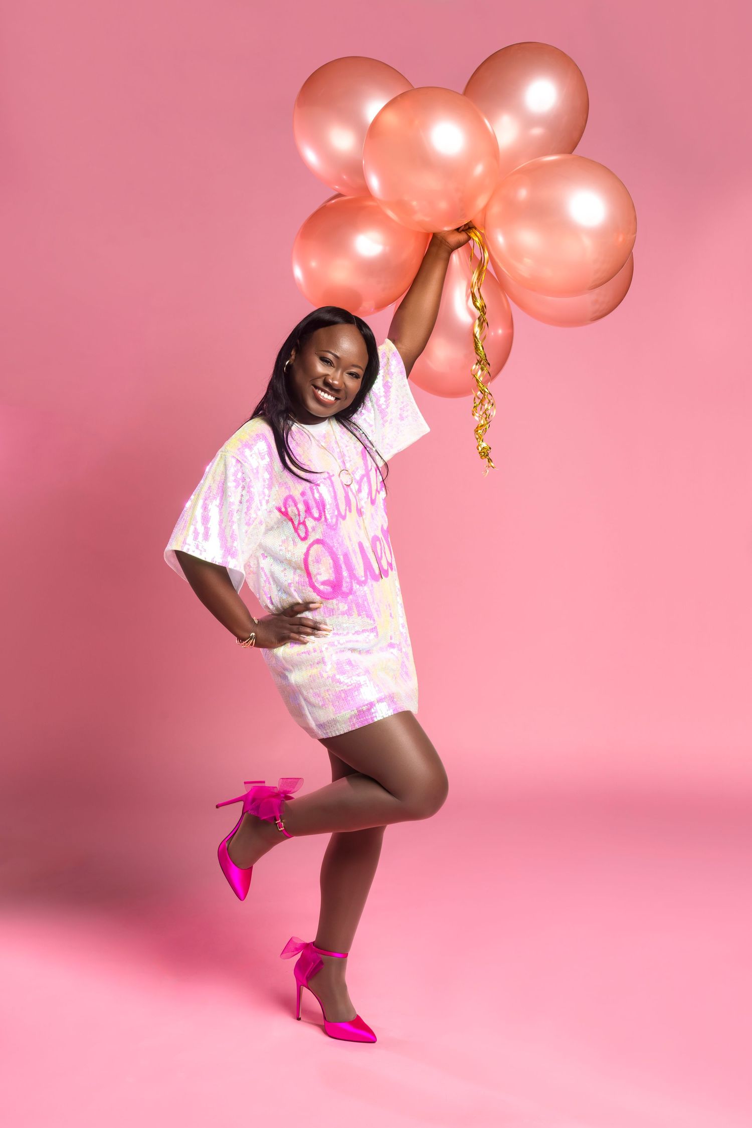 A joyful figure in a pink and white t-shirt poses with rose gold balloons against a pink backdrop wearing bright pink heels.