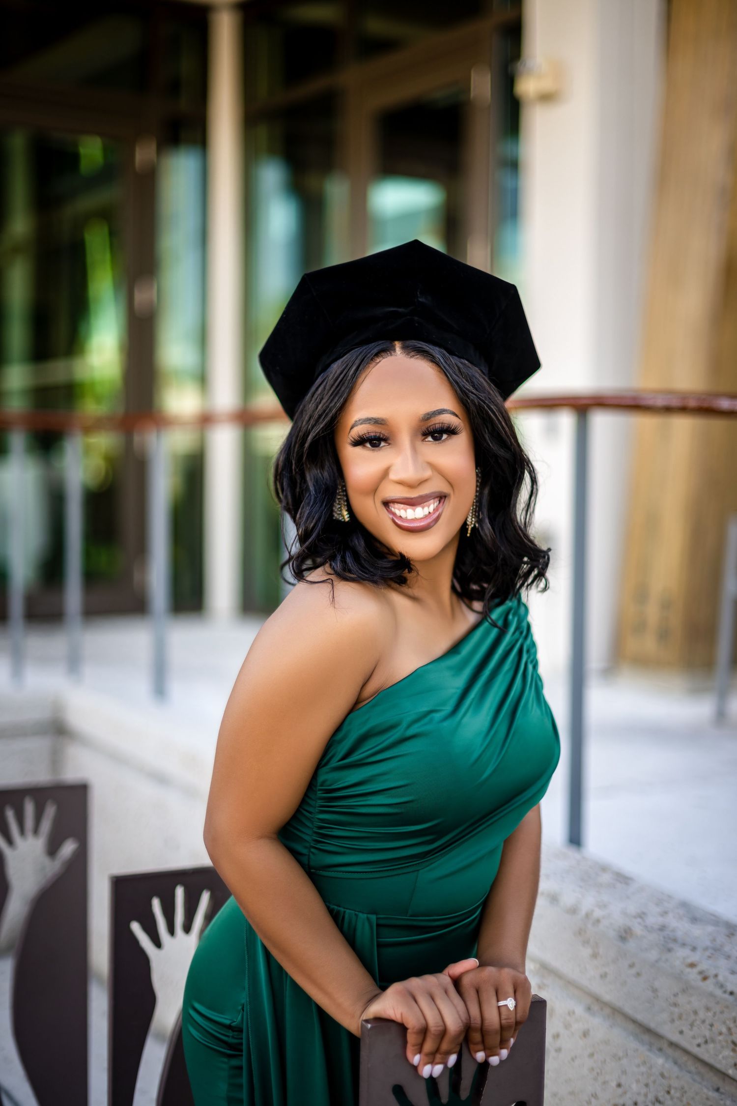 Graduate in emerald green one-shoulder dress and black cap poses with bright smile outside building.