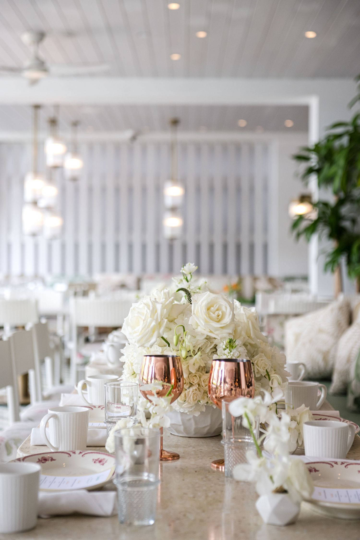 Elegant white floral centerpieces with roses in copper vases adorn tables at an upscale wedding reception venue.