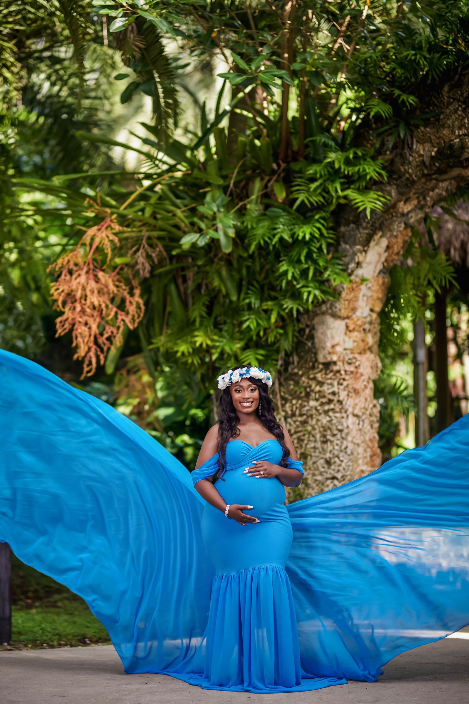A flowing bright blue maternity gown billows dramatically in a lush garden setting during a photo session.