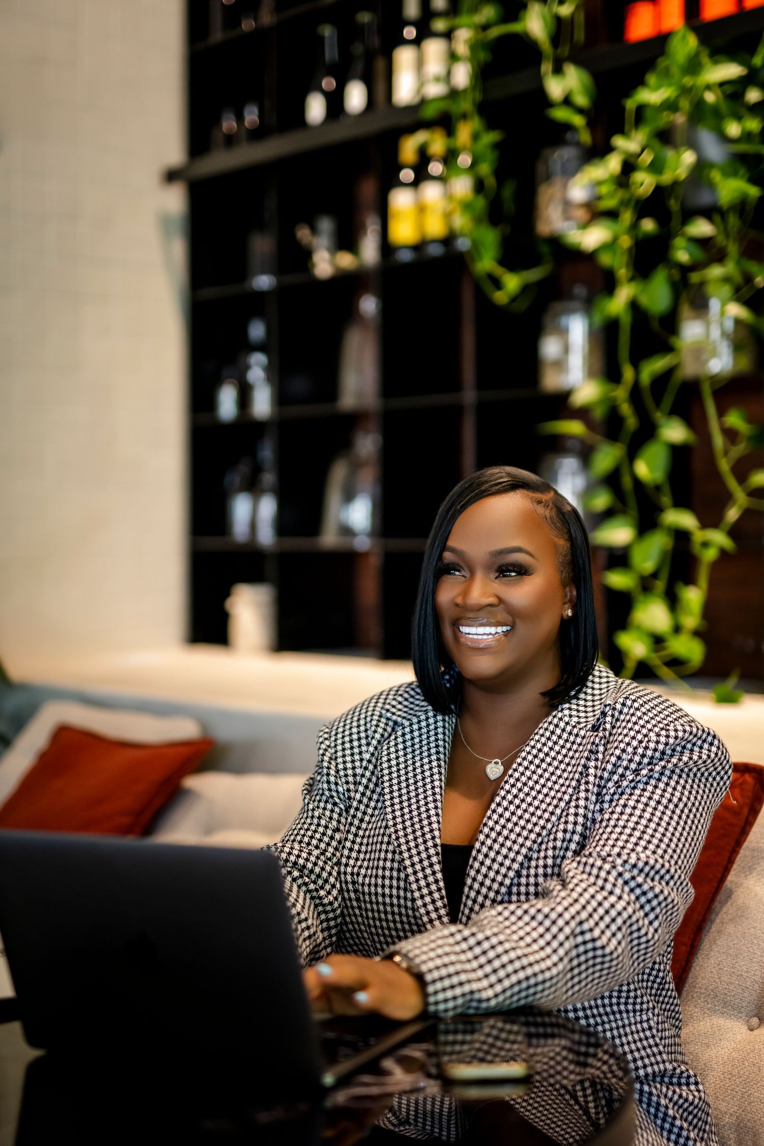 Professional in black and white gingham blazer works on laptop in modern office setting with wine bottles in background.