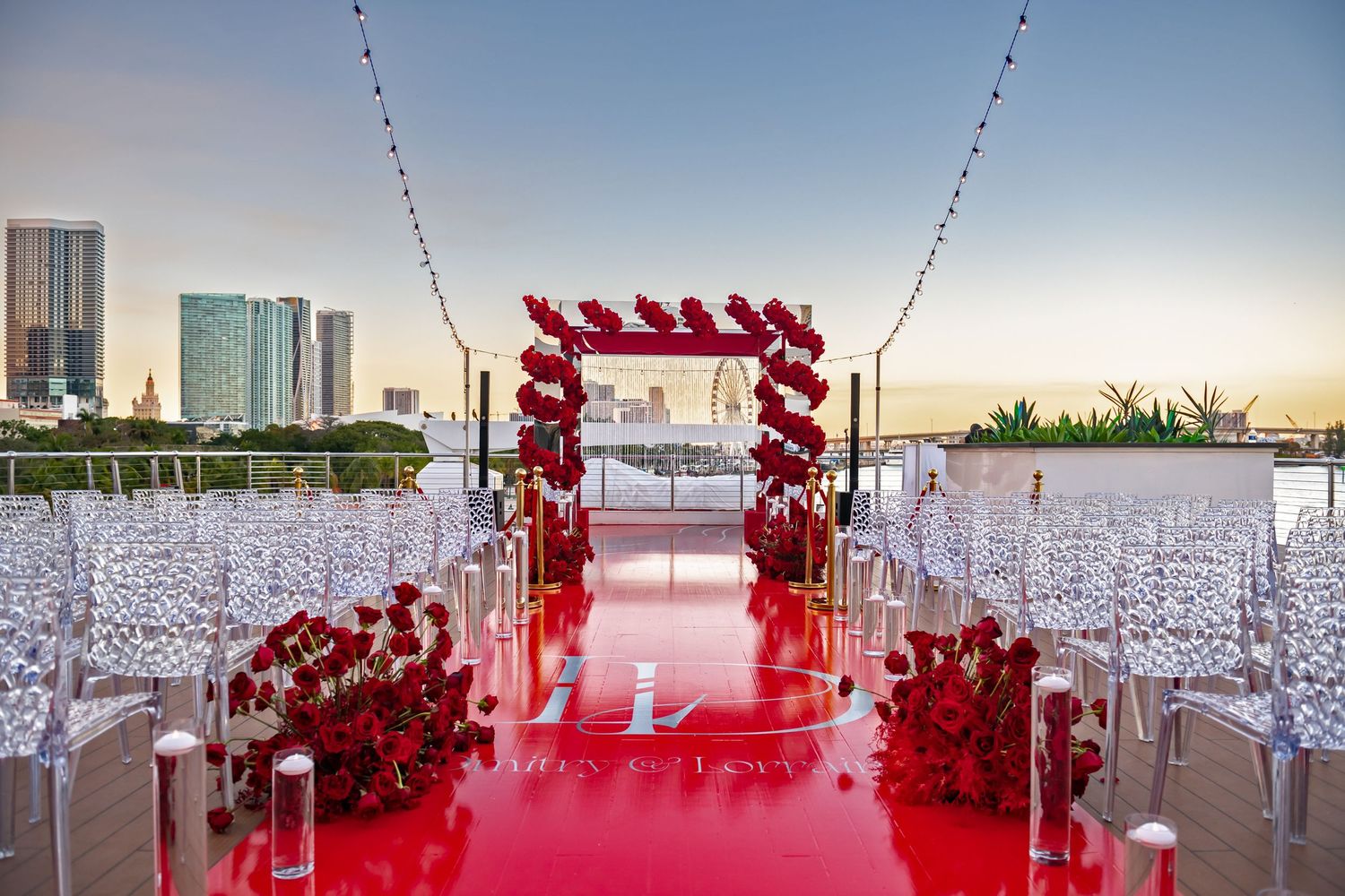 Elegant outdoor wedding ceremony setup with red carpet aisle, floral arch and white chairs overlooking city skyline at sunset.