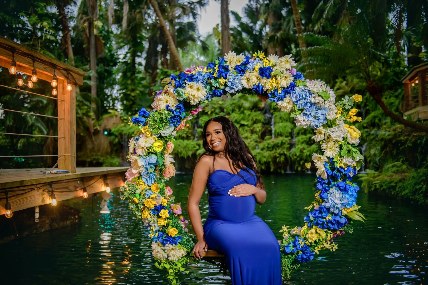 A person in a royal blue dress stands near a pond surrounded by lush greenery and floral decorations.