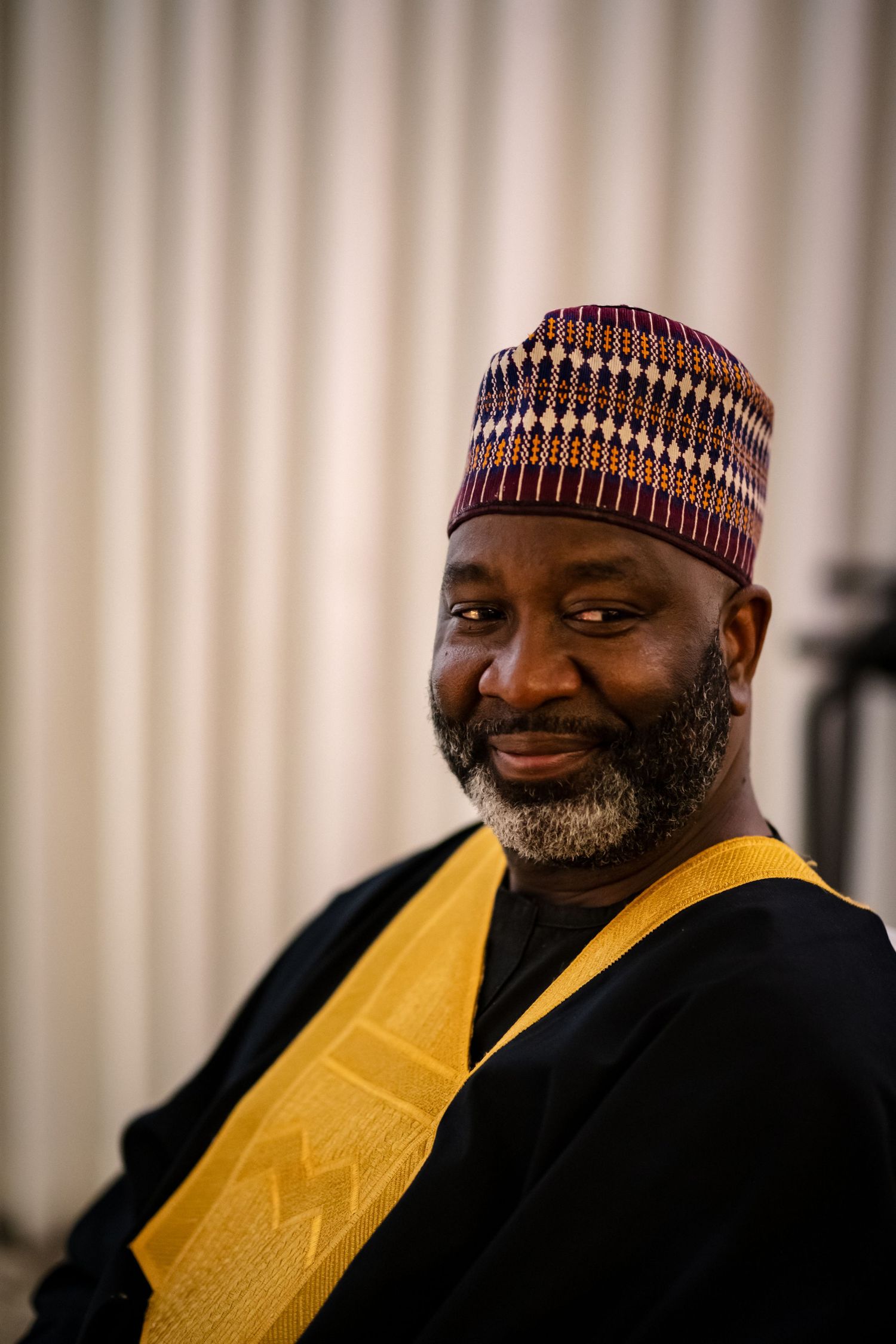 A person wearing a striped hat and yellow traditional garment smiles warmly at the camera.