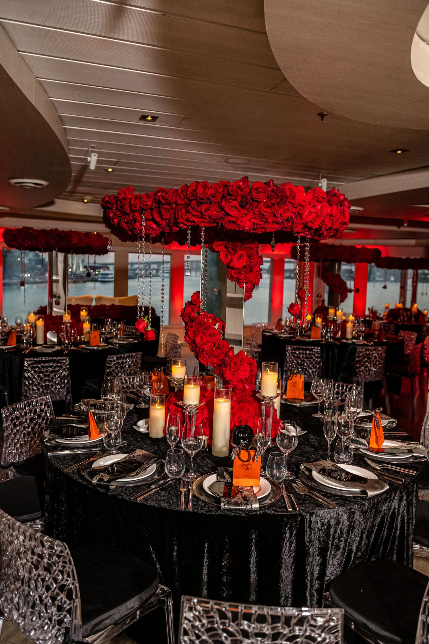 Modern restaurant interior featuring black-draped tables adorned with red floral arrangements and crystal chandeliers.