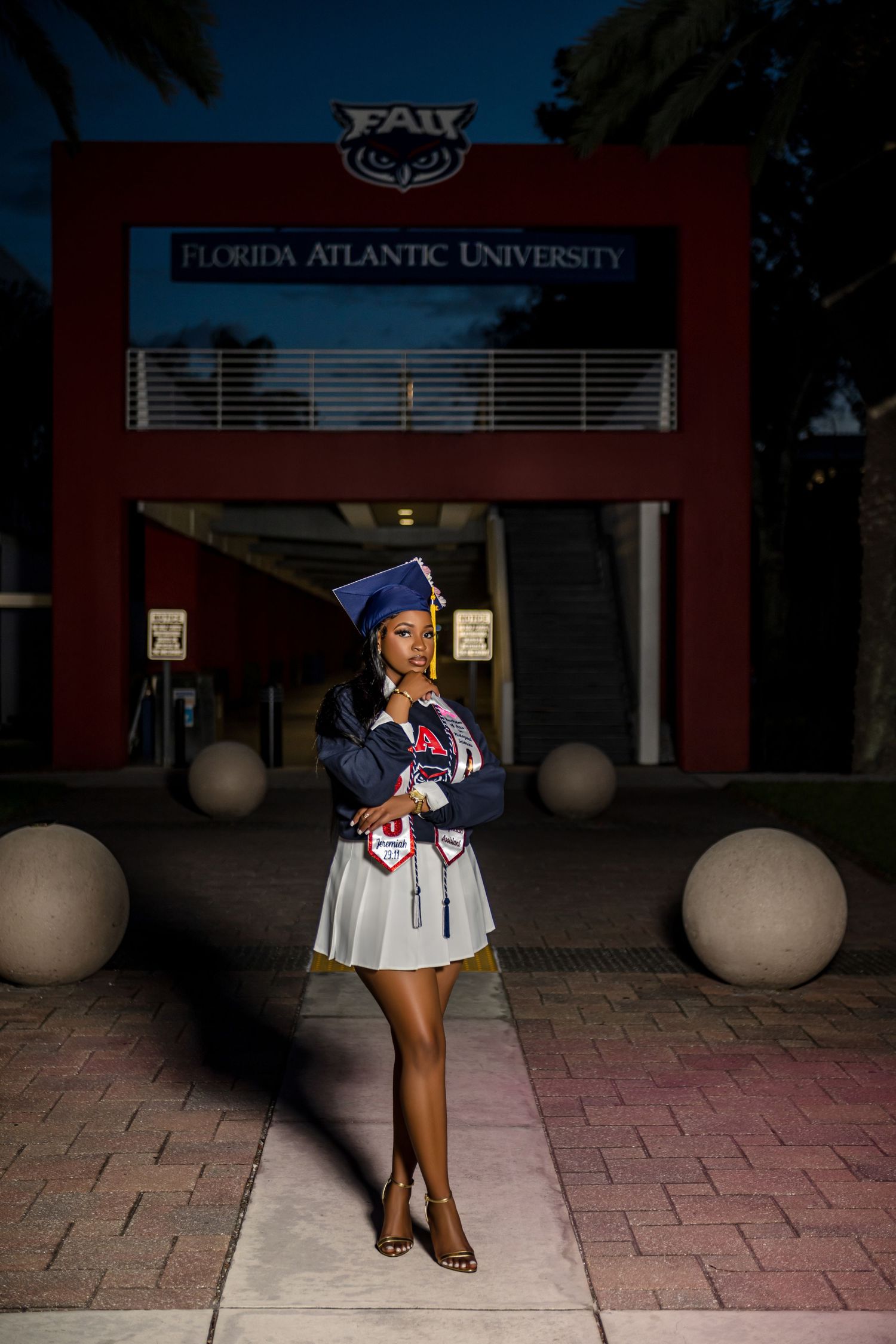 Graduate in blue cap and white dress poses at night in front of Florida Atlantic University entrance sign.