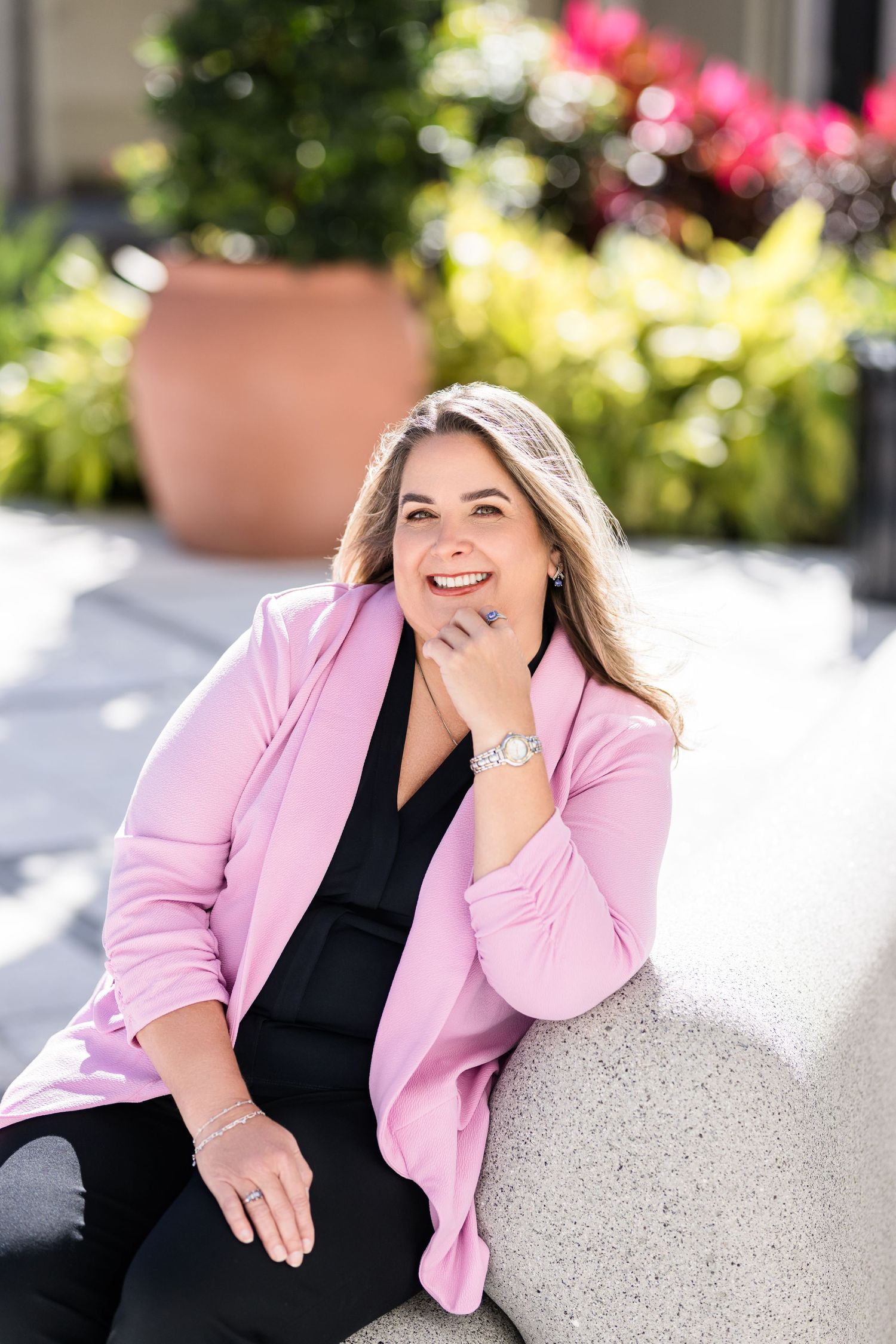 A professional portrait in a pink blazer and black top sitting outdoors with greenery and flowers in the background.