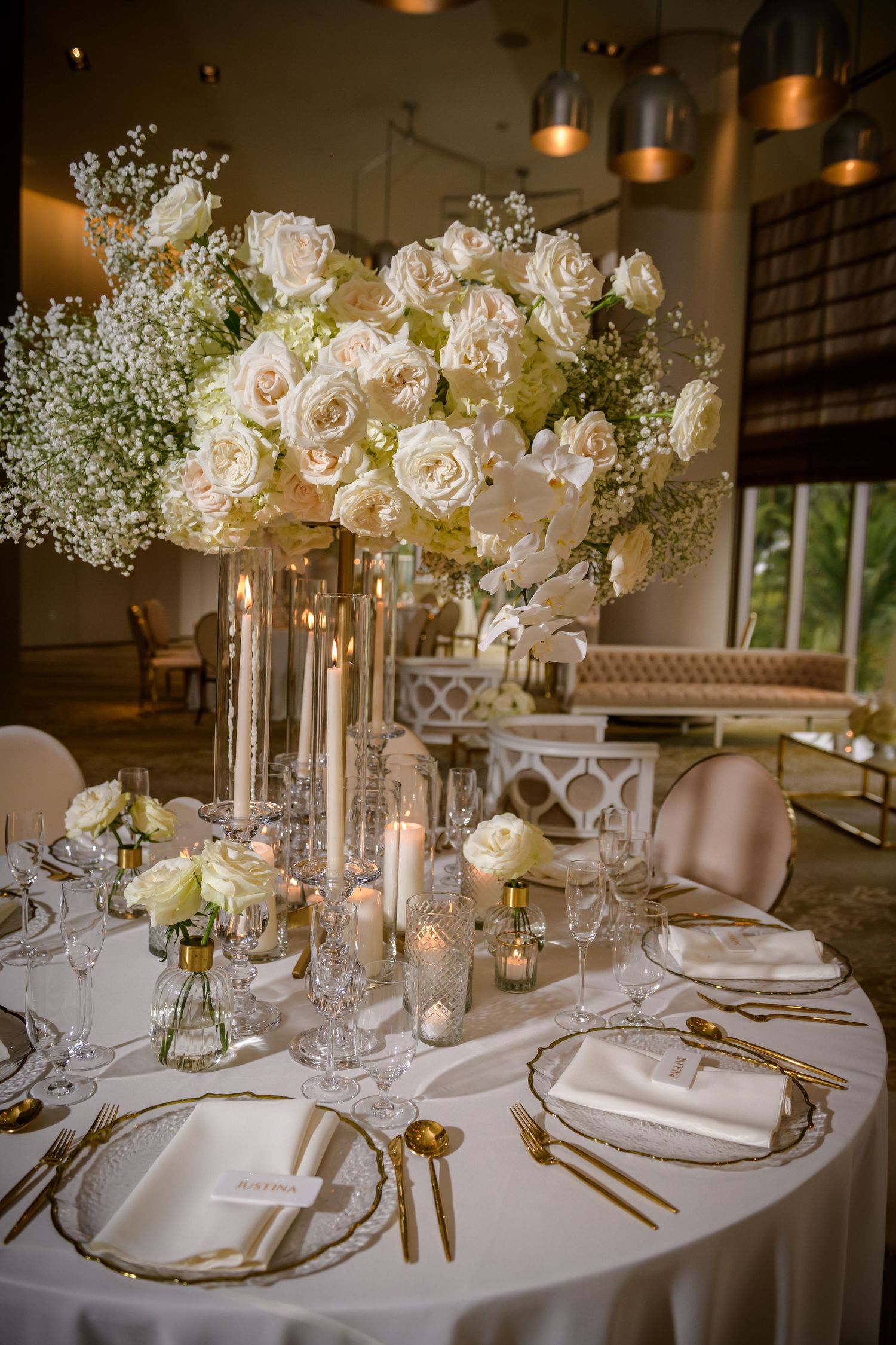 Elegant wedding table settings with white roses, crystal vases, and glowing candles in a luxurious indoor venue.