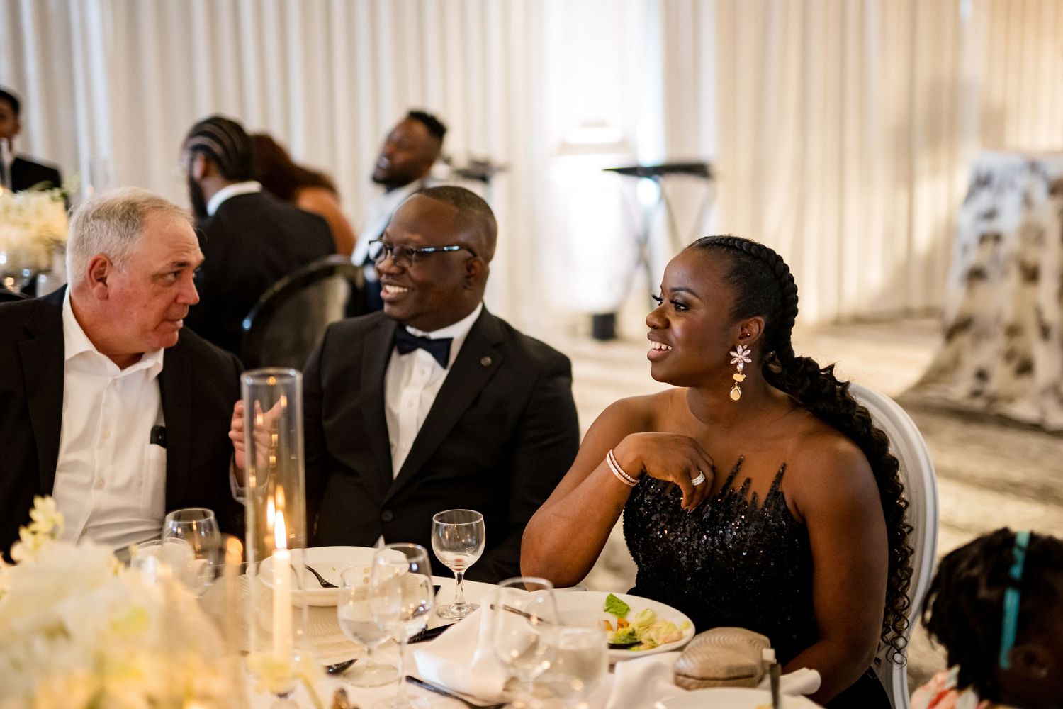 Wedding guests seated at elegant reception tables engage in conversation during a formal dinner celebration.