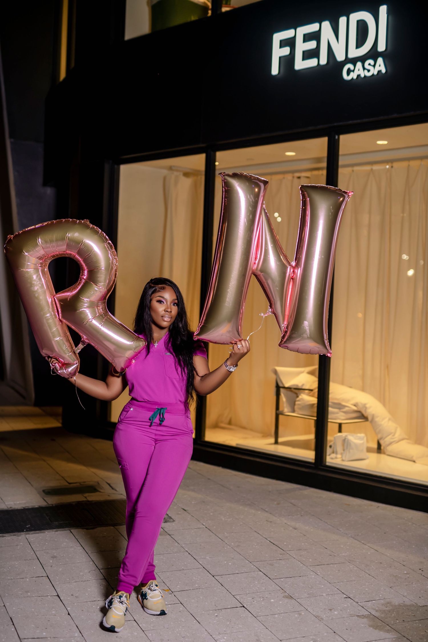Woman in bright pink dress poses playfully with balloon letters outside Fendi Casa store at night.