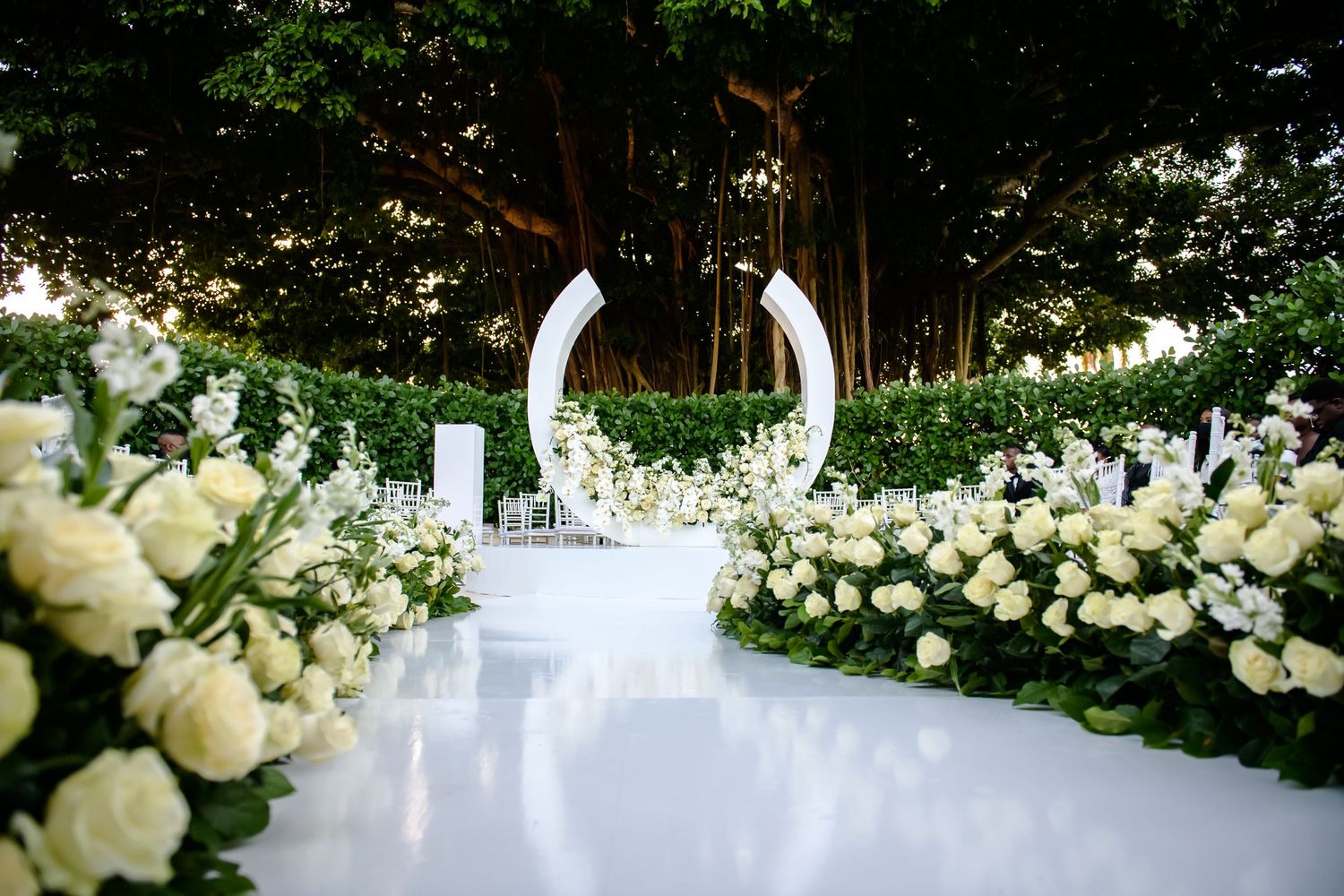 Outdoor wedding ceremony setup with white floral arch and rose-lined aisle.