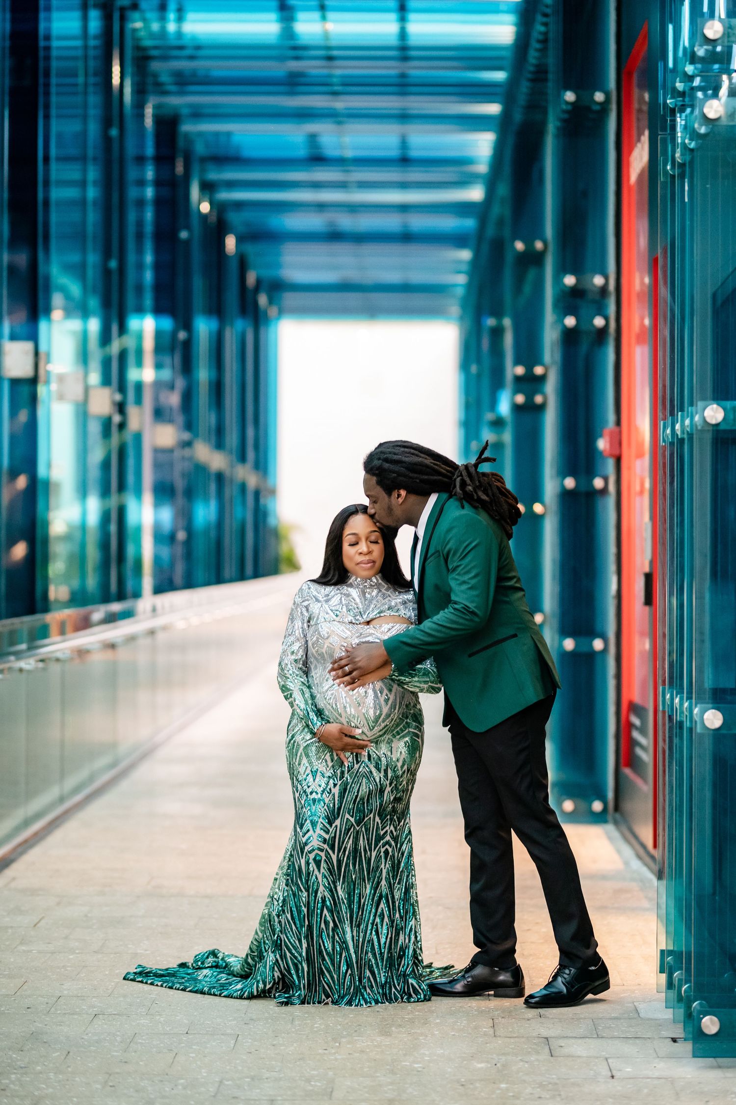 A romantic couple shares tender moments in a modern glass corridor with turquoise lighting and reflective surfaces.