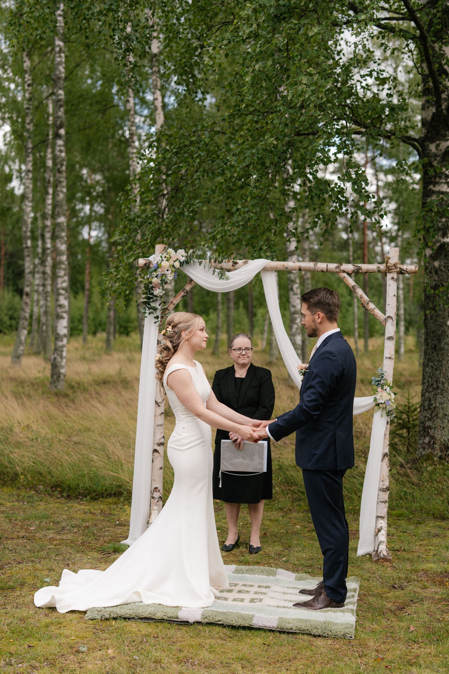 Wedding couple exchanges vows in front of an officiant beneath a white draped arch in a birch grove.