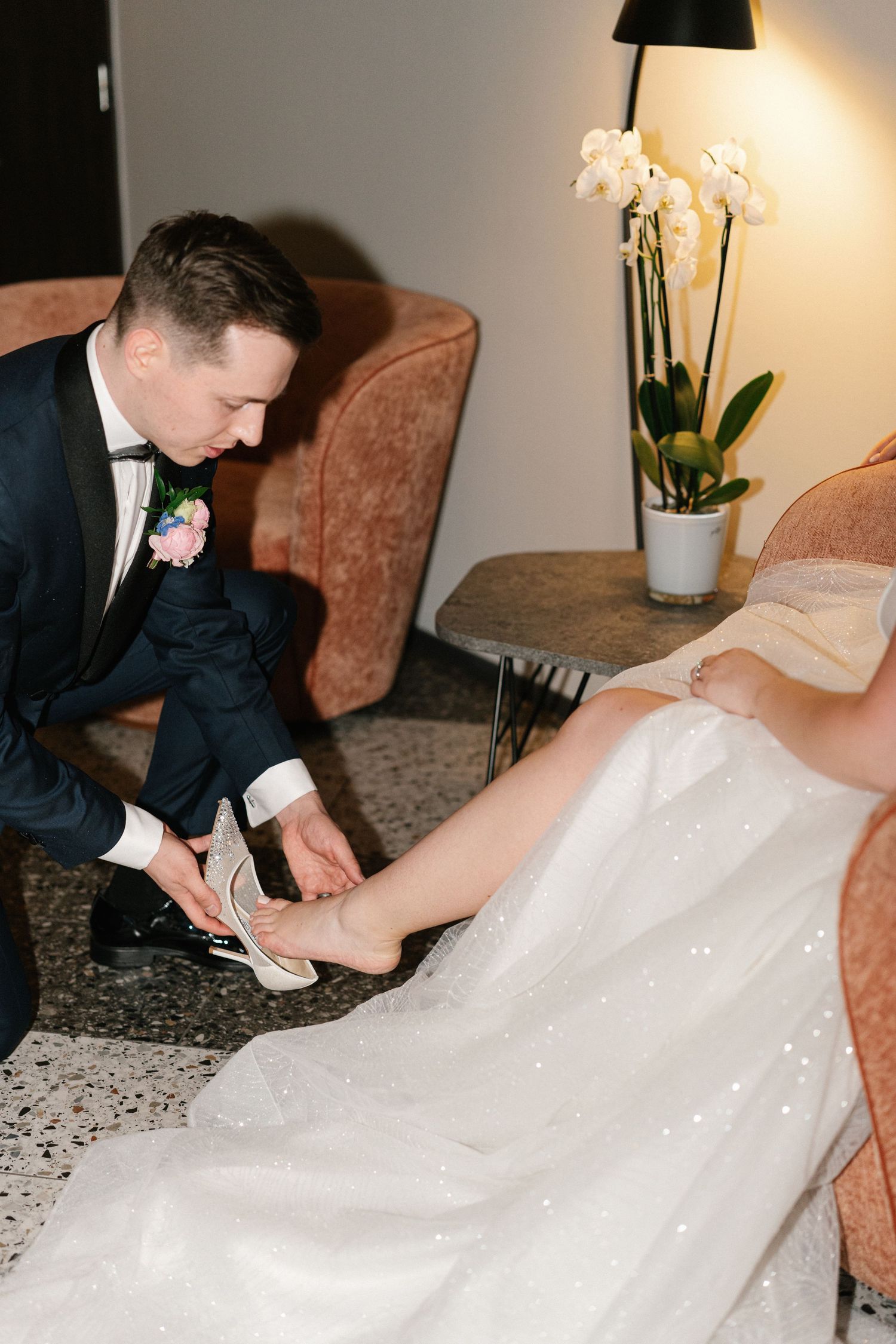 A person in a tuxedo helps adjust the flowing white wedding dress train on the floor.