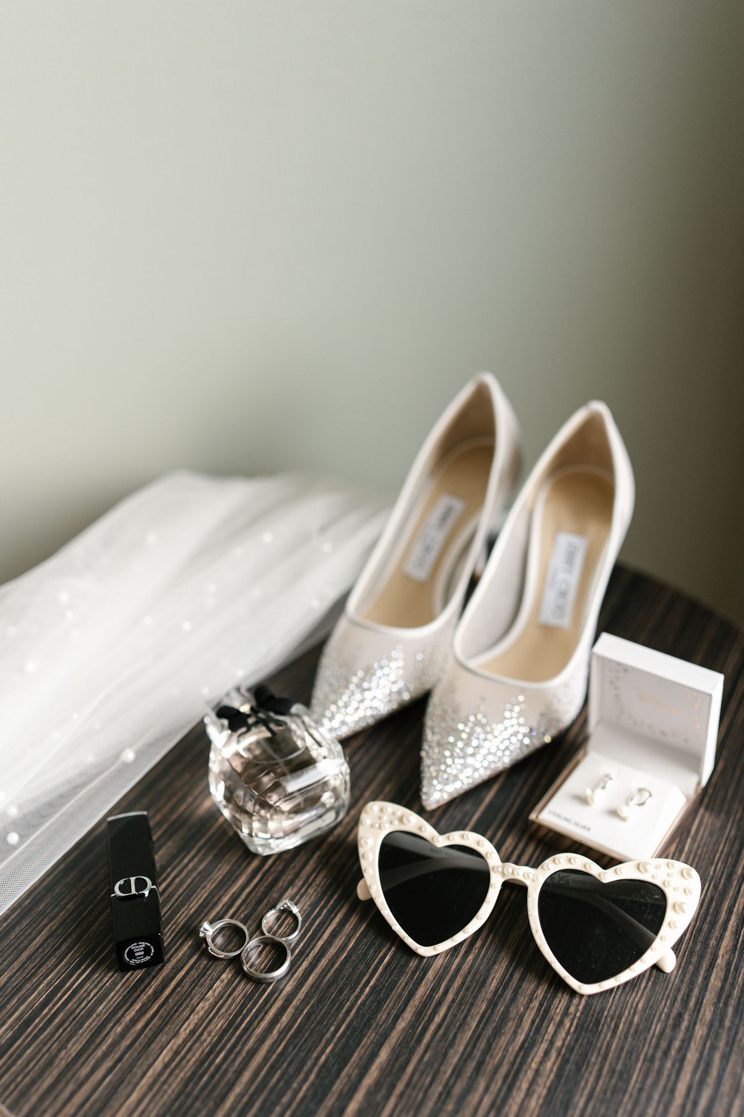 Elegant bridal shoes, heart-shaped sunglasses, and wedding rings arranged on dark wood surface.