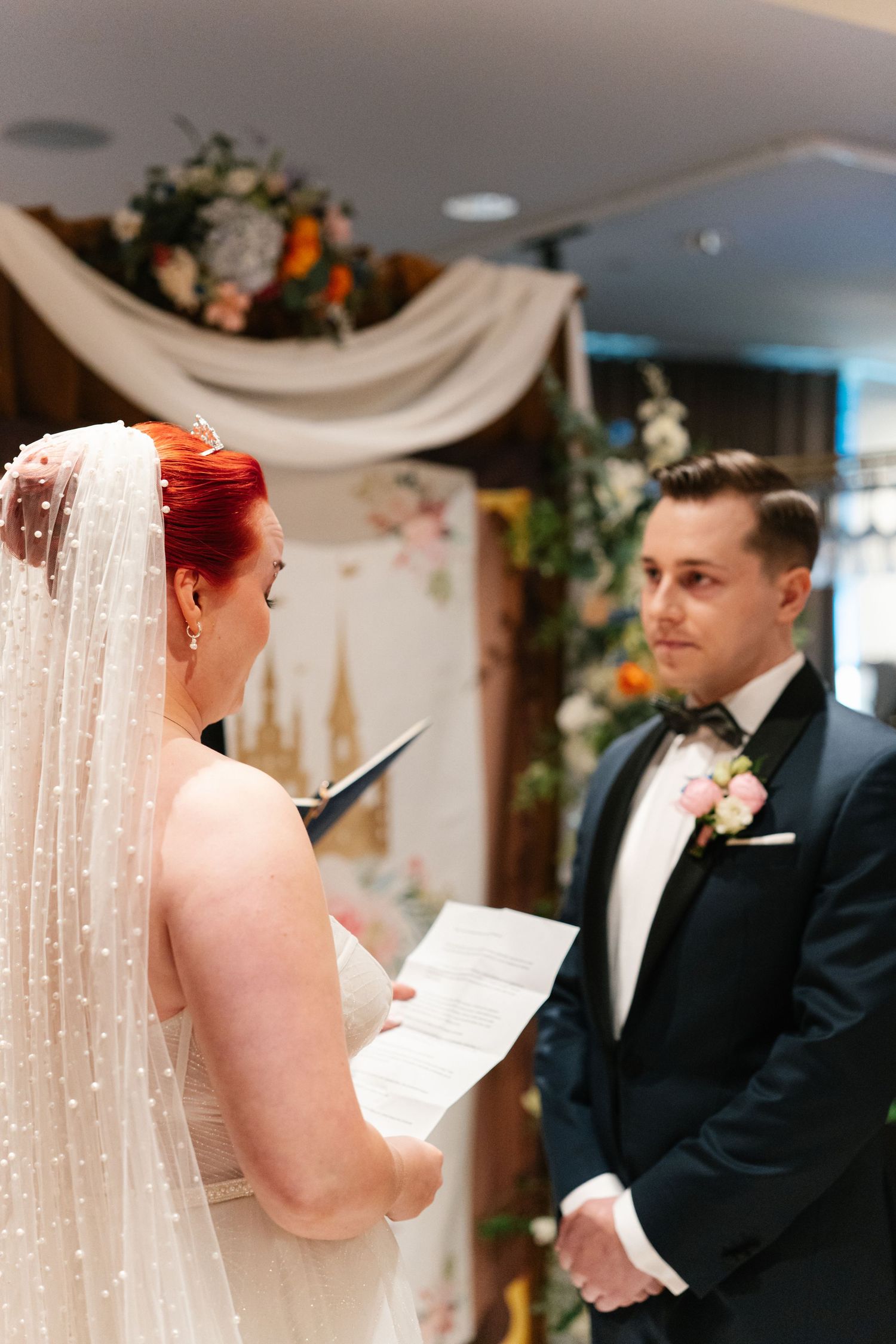 Wedding ceremony with couple reading vows in front of decorative floral backdrop.