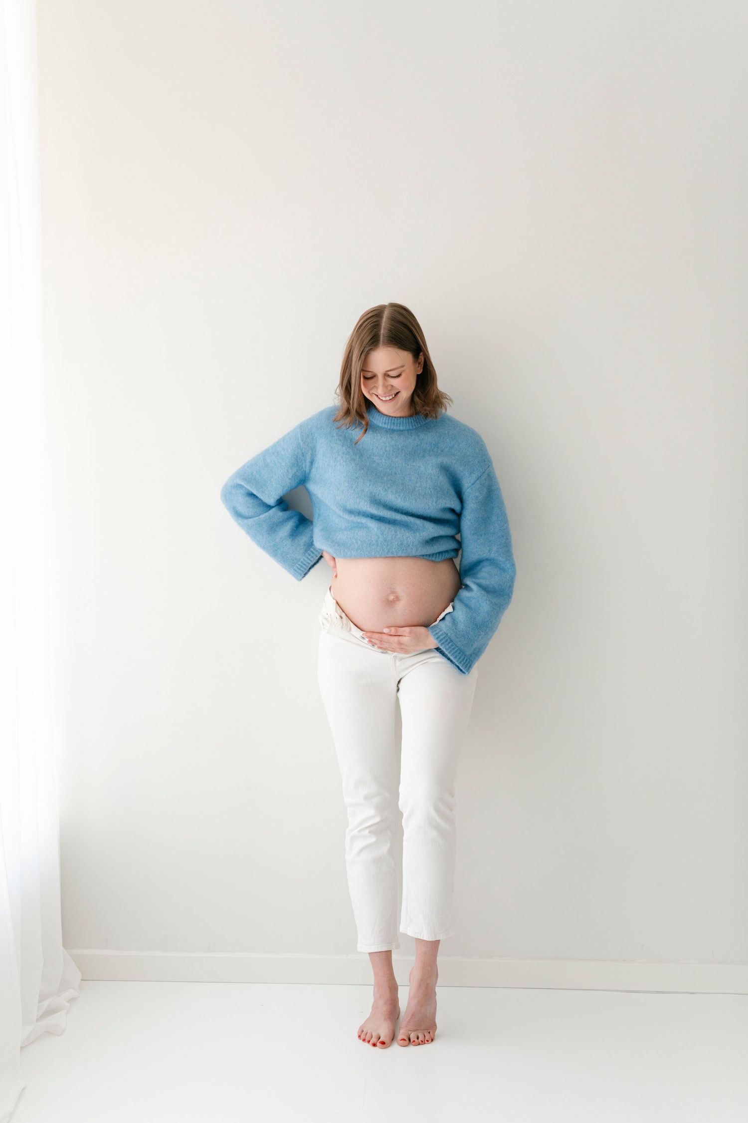 Maternity photos with woman in blue sweater and white pants against minimalist background.