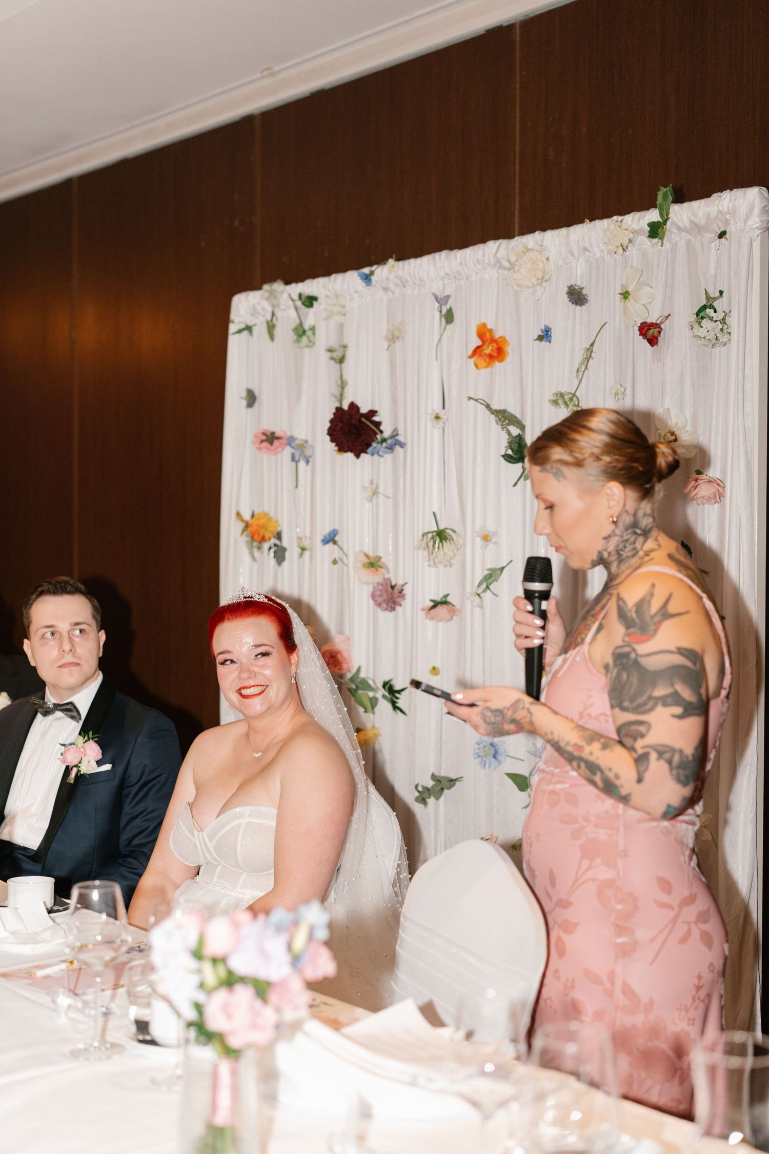 Wedding reception guests in formal attire seated at decorated tables with floral arrangements.