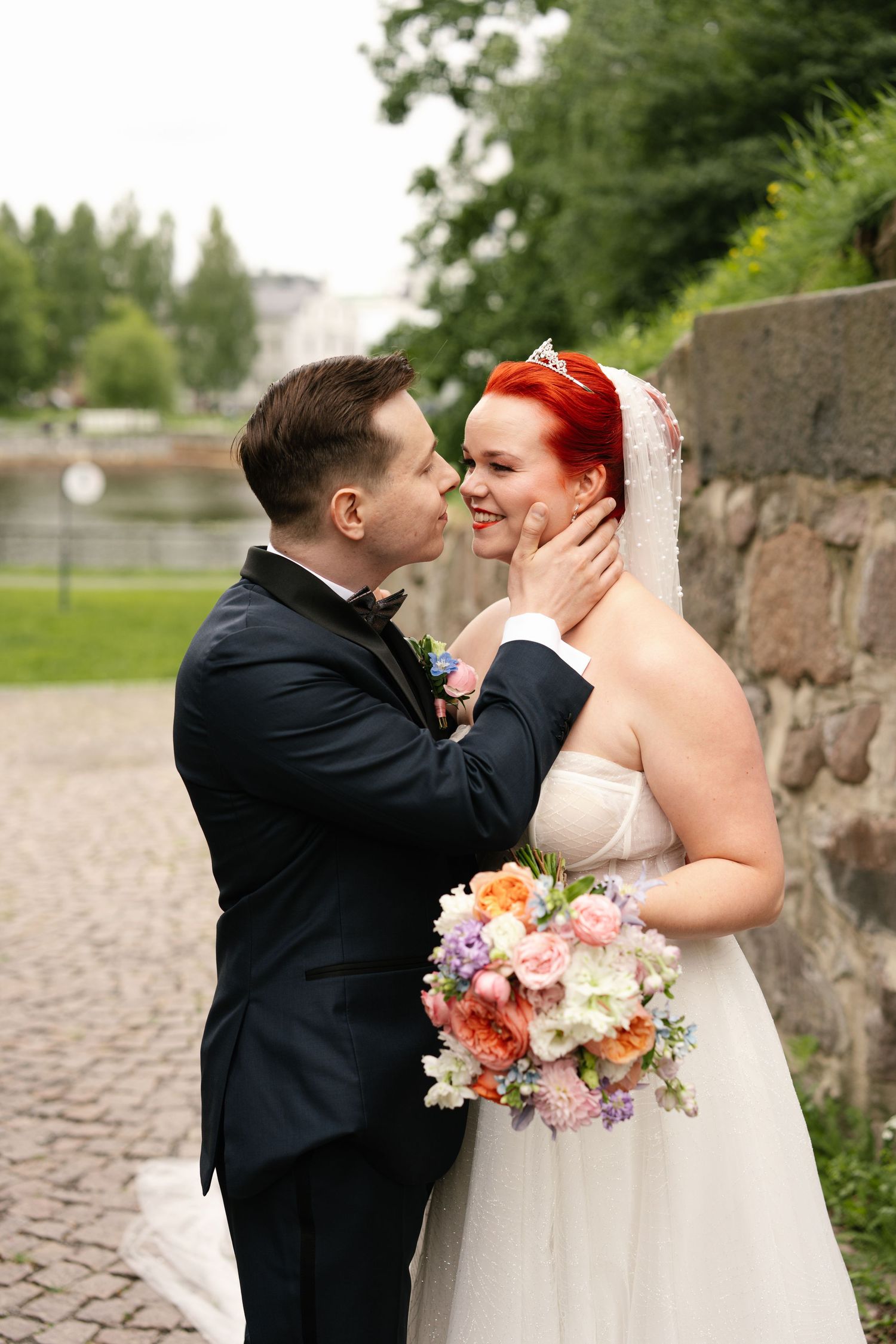 Wedding couple shares intimate moments against stone wall with vibrant floral bouquet.