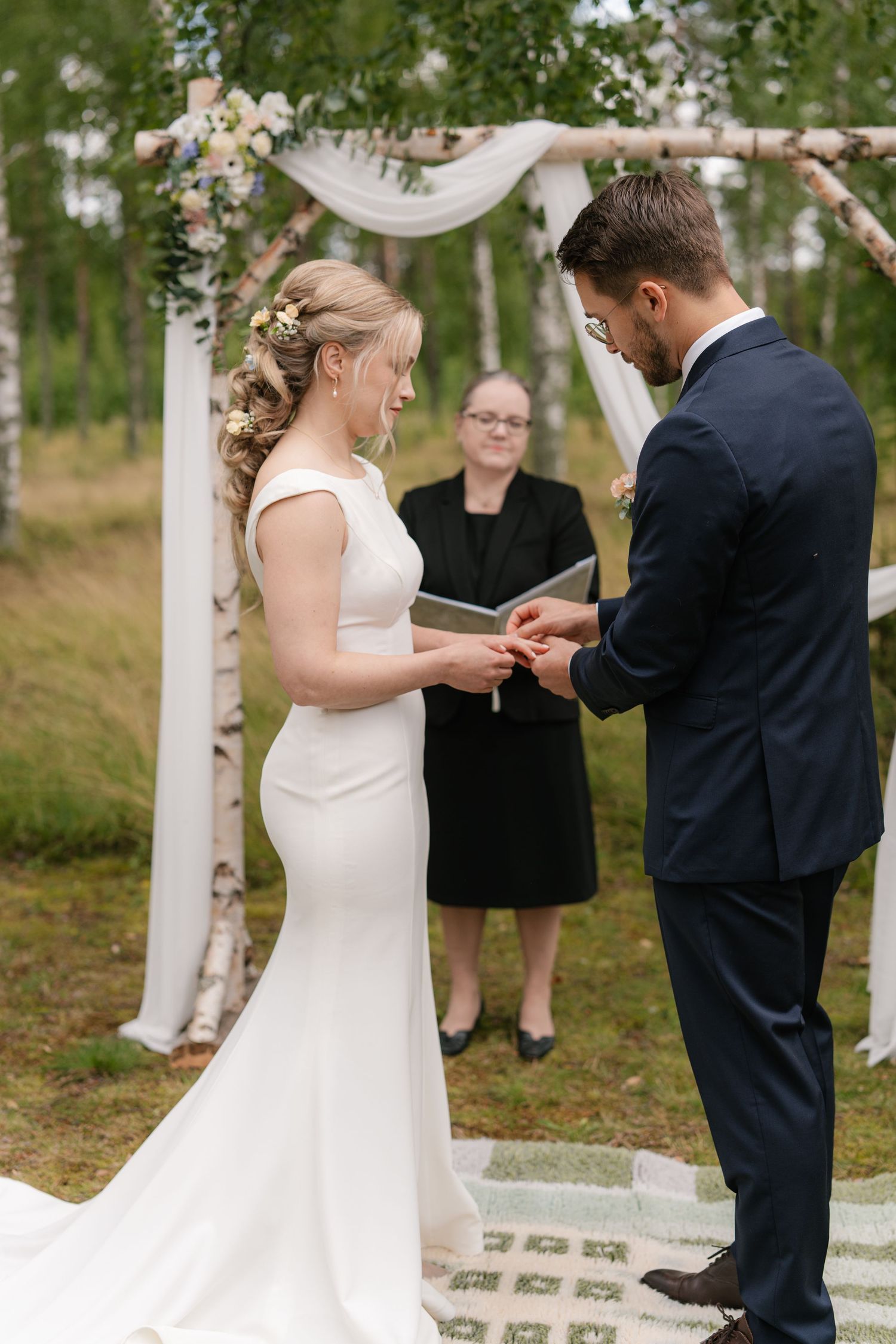Wedding couple participates in ring exchange ceremony under decorated arch in woodland setting.