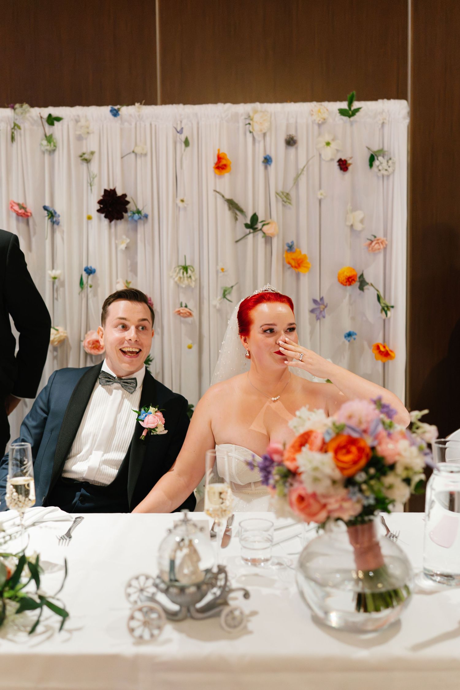 Wedding reception table decorated with hanging floral elements and white draping in the background.