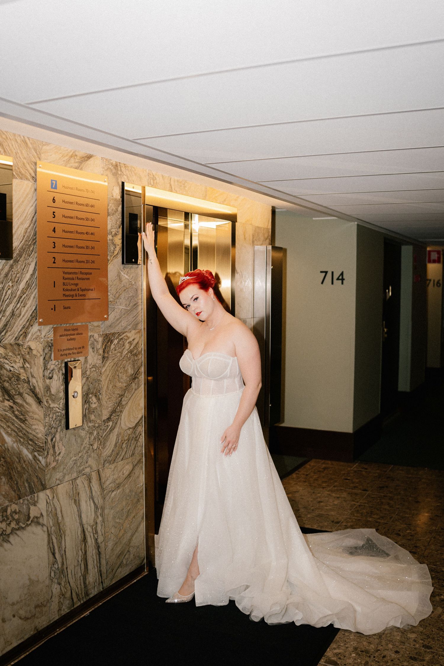 A flowing white wedding gown with a long train is displayed in an elegant hotel hallway with marble walls.