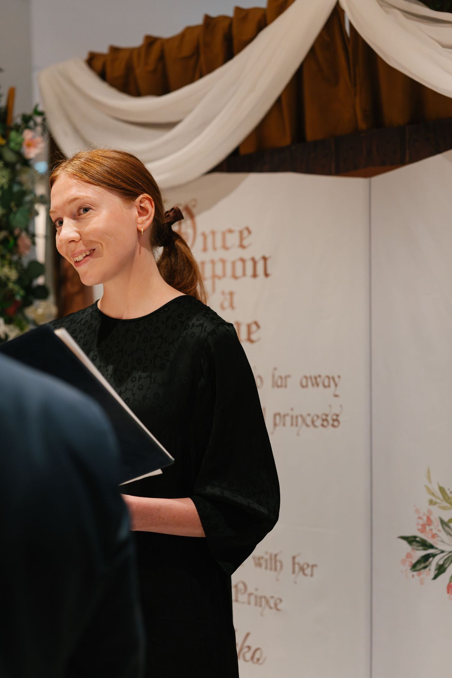 Wedding officiant in black attire conducting the ceremony against a decorated backdrop.