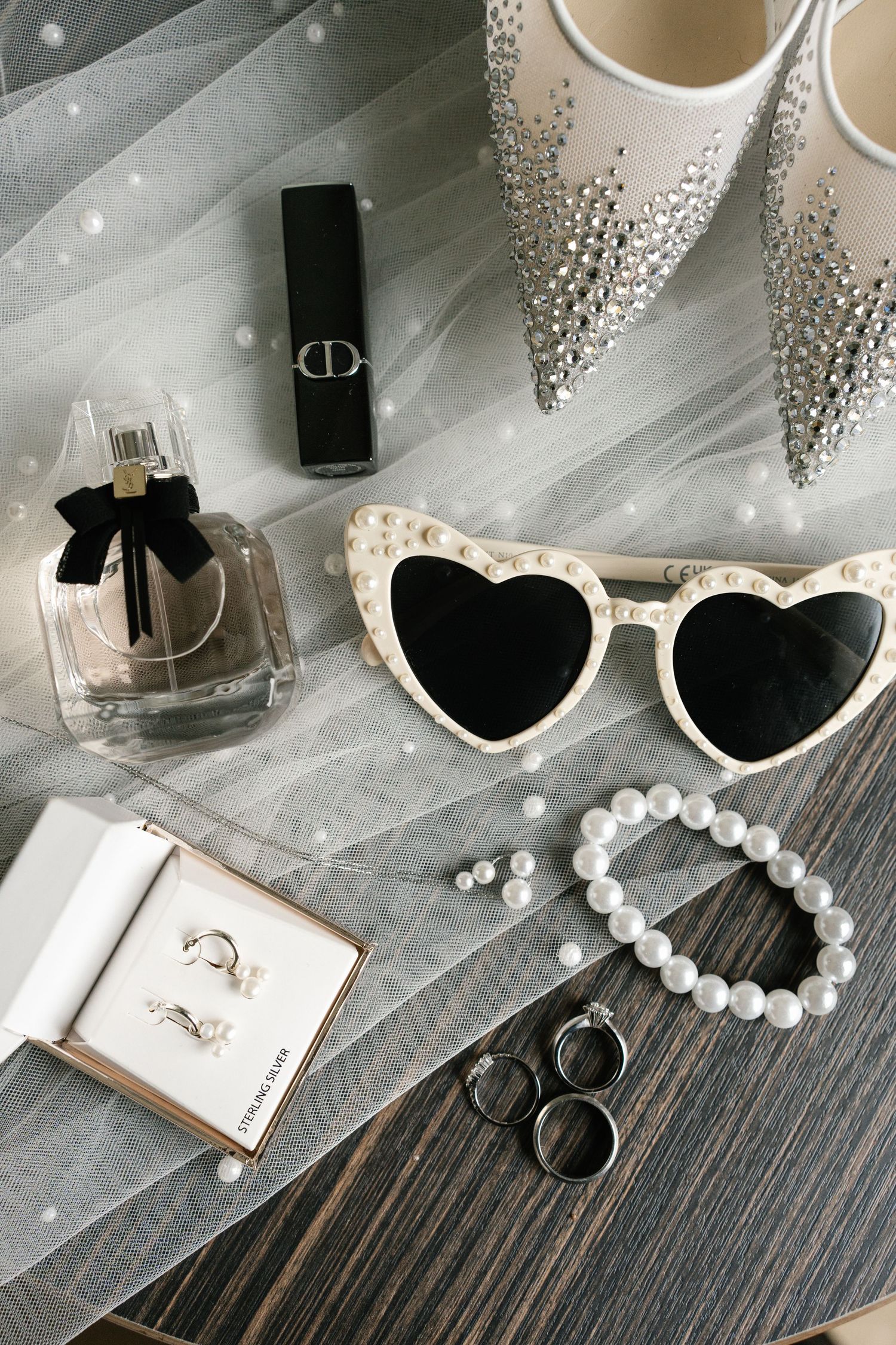 Wedding accessories including heart sunglasses, pearl bracelet, rings and perfume on wooden surface.