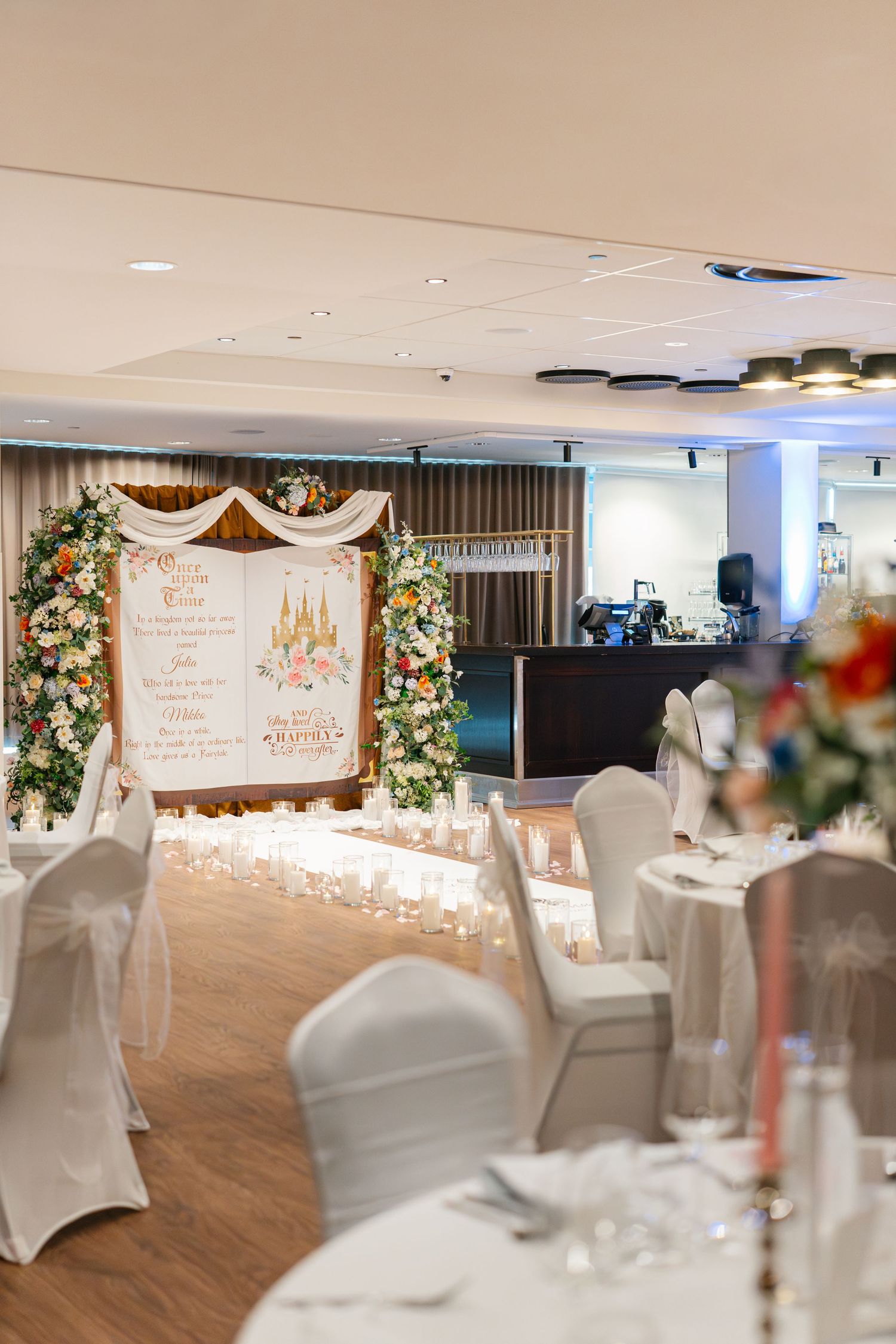 Wedding reception venue with white-covered chairs and floral decorations along the walls.