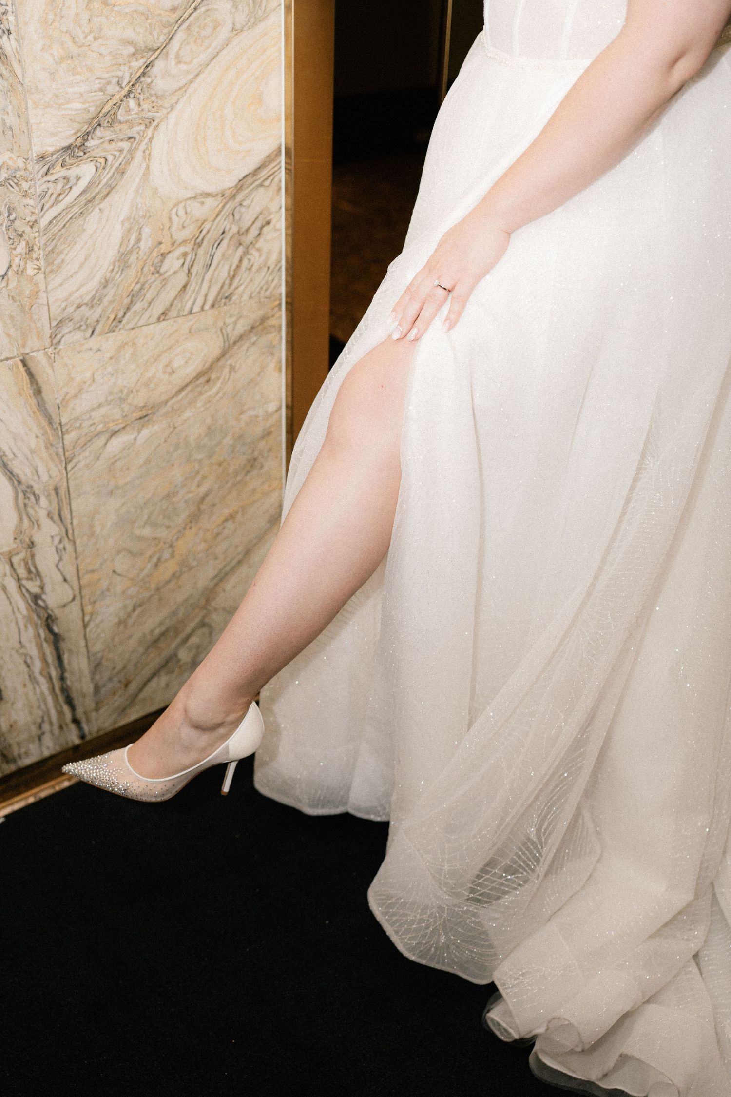 The hem of a flowing white wedding gown cascades gracefully against a marble wall.