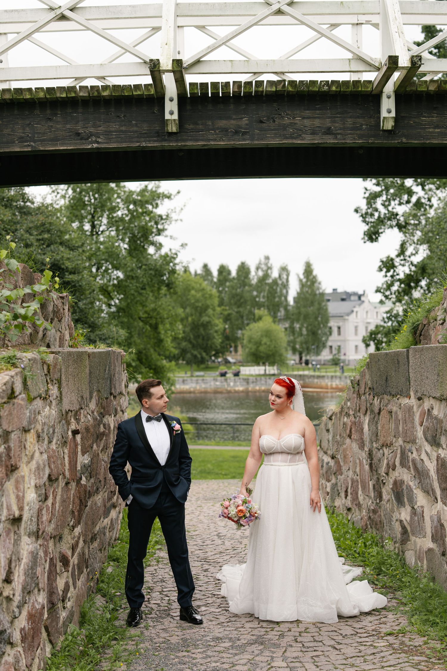 Wedding couple poses along rustic stone wall and wooden bridge in outdoor setting.
