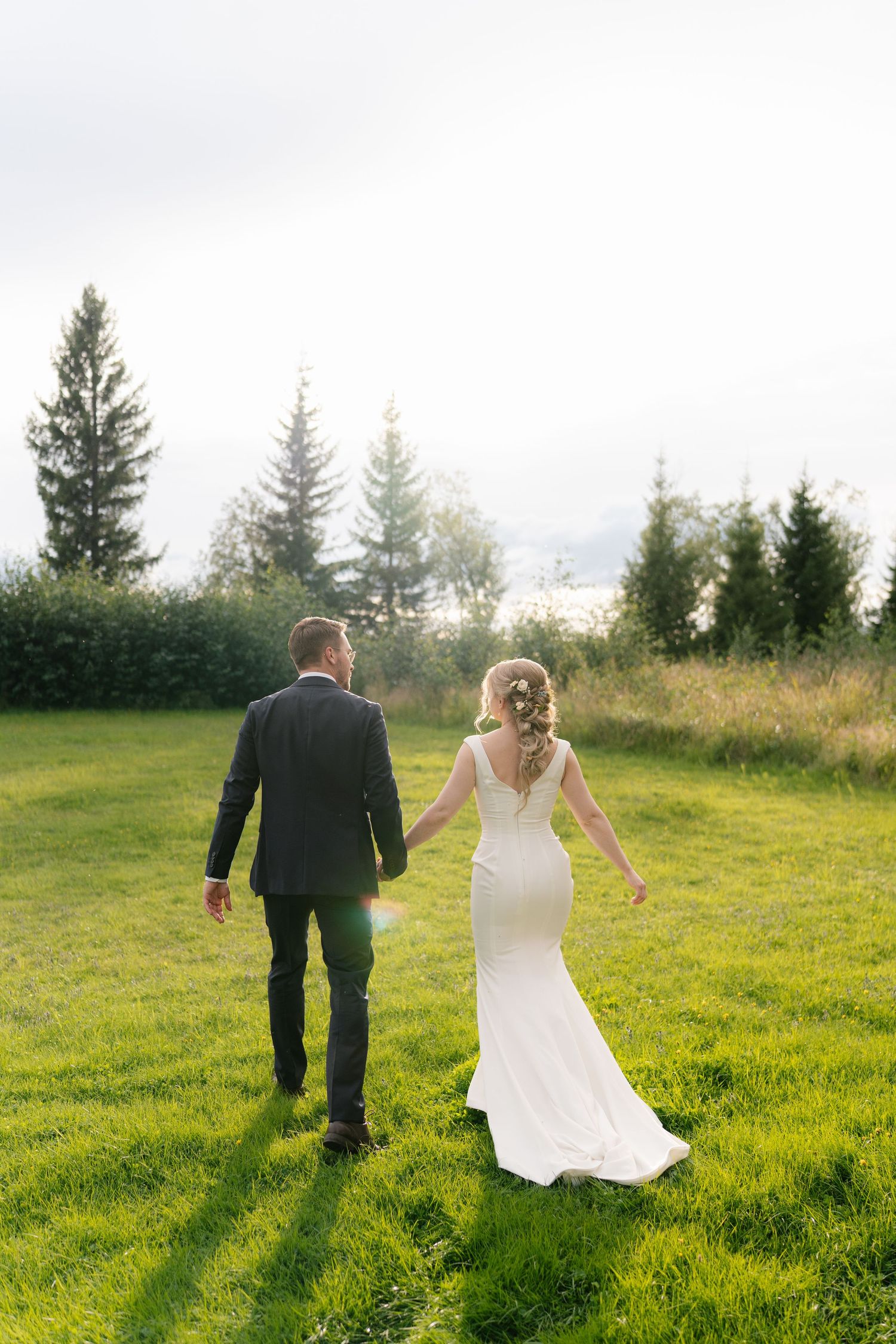 Newlyweds walk hand in hand through a lush green field with pine trees in the background.