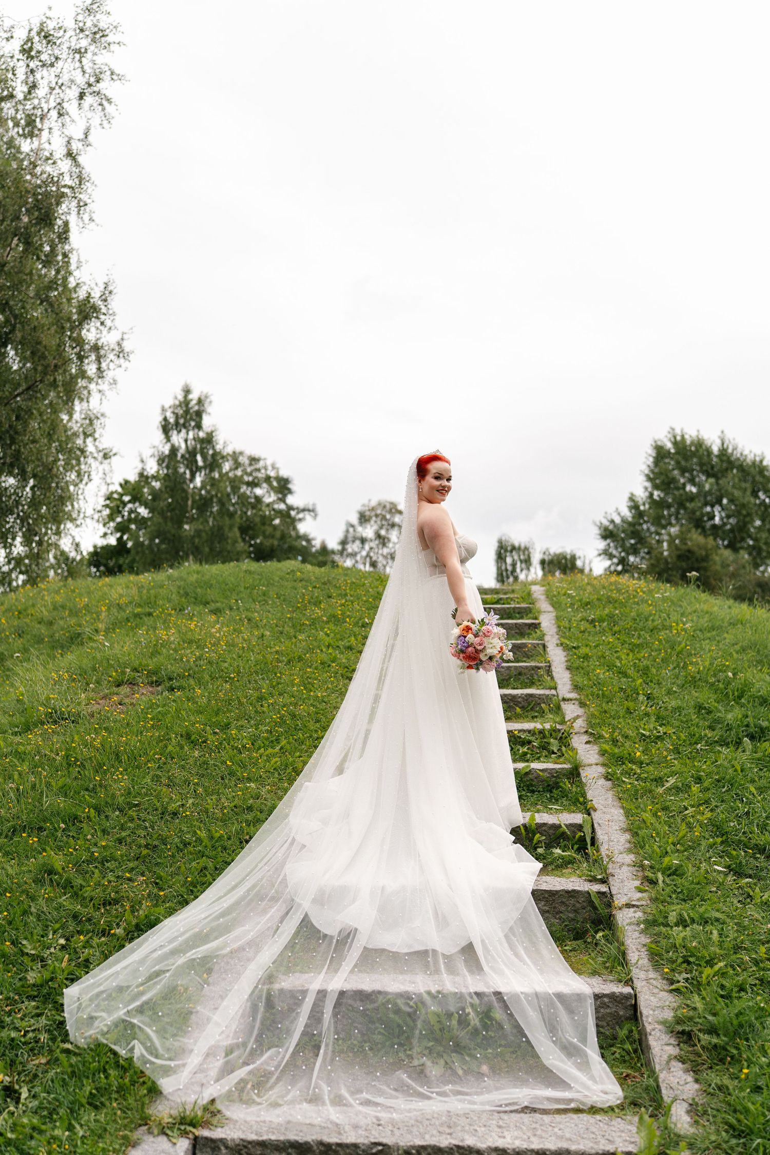 Flowing white wedding dress and veil trailing down stone steps in a garden setting.