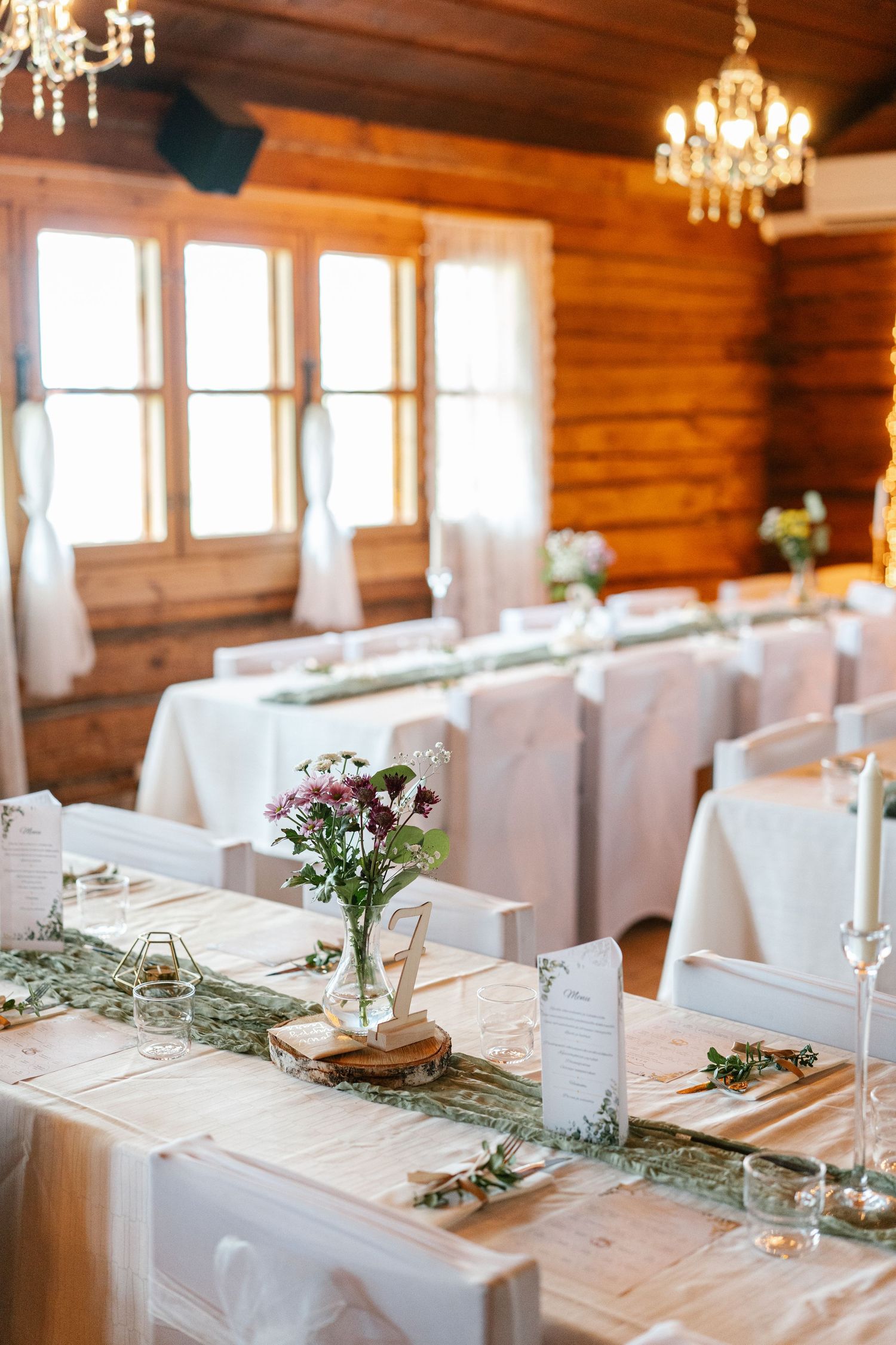 Wedding reception tables decorated with greenery runners and crystal chandeliers in log cabin setting.