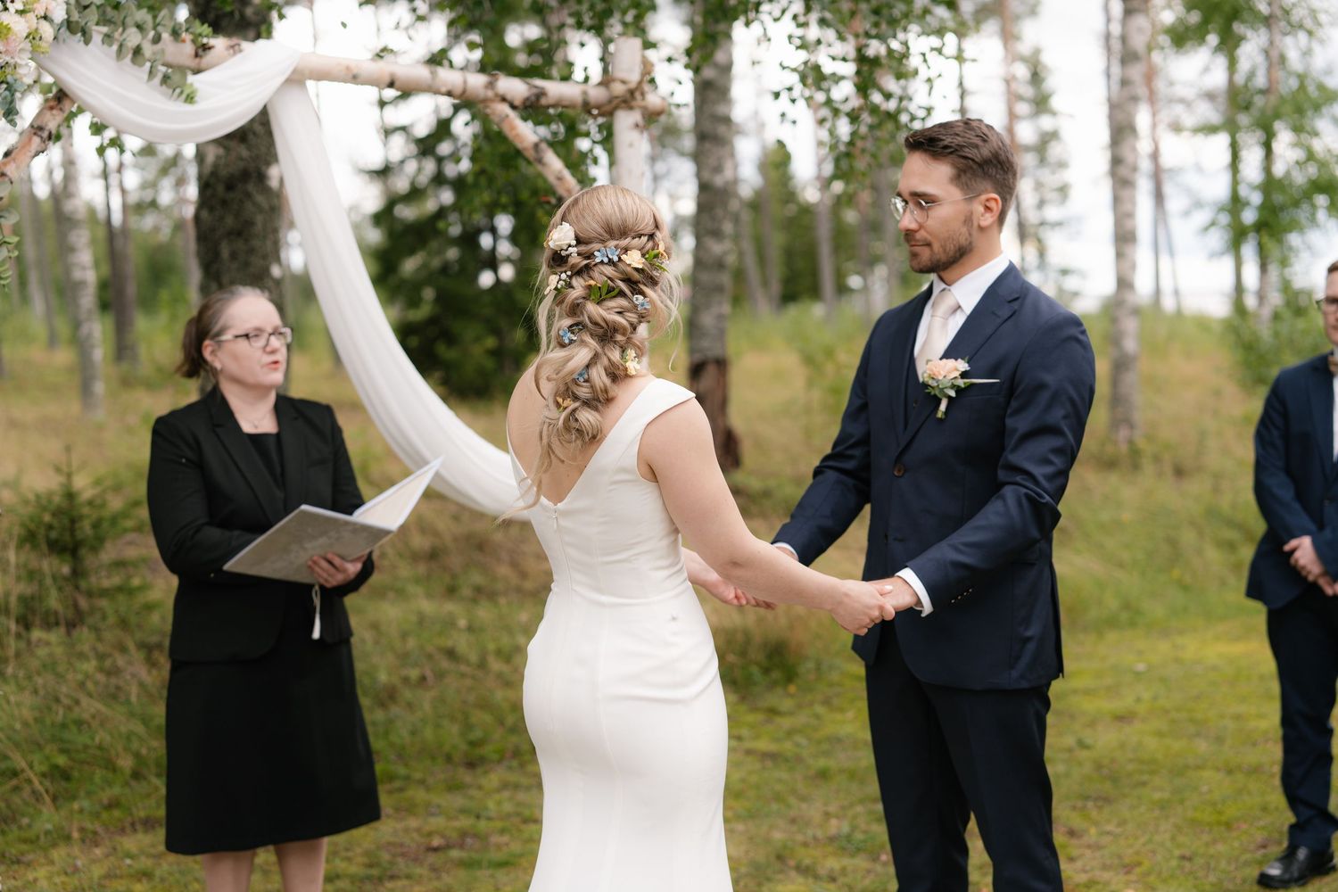 A wedding ceremony takes place in a serene birch forest with a white fabric arch and floral decorations.